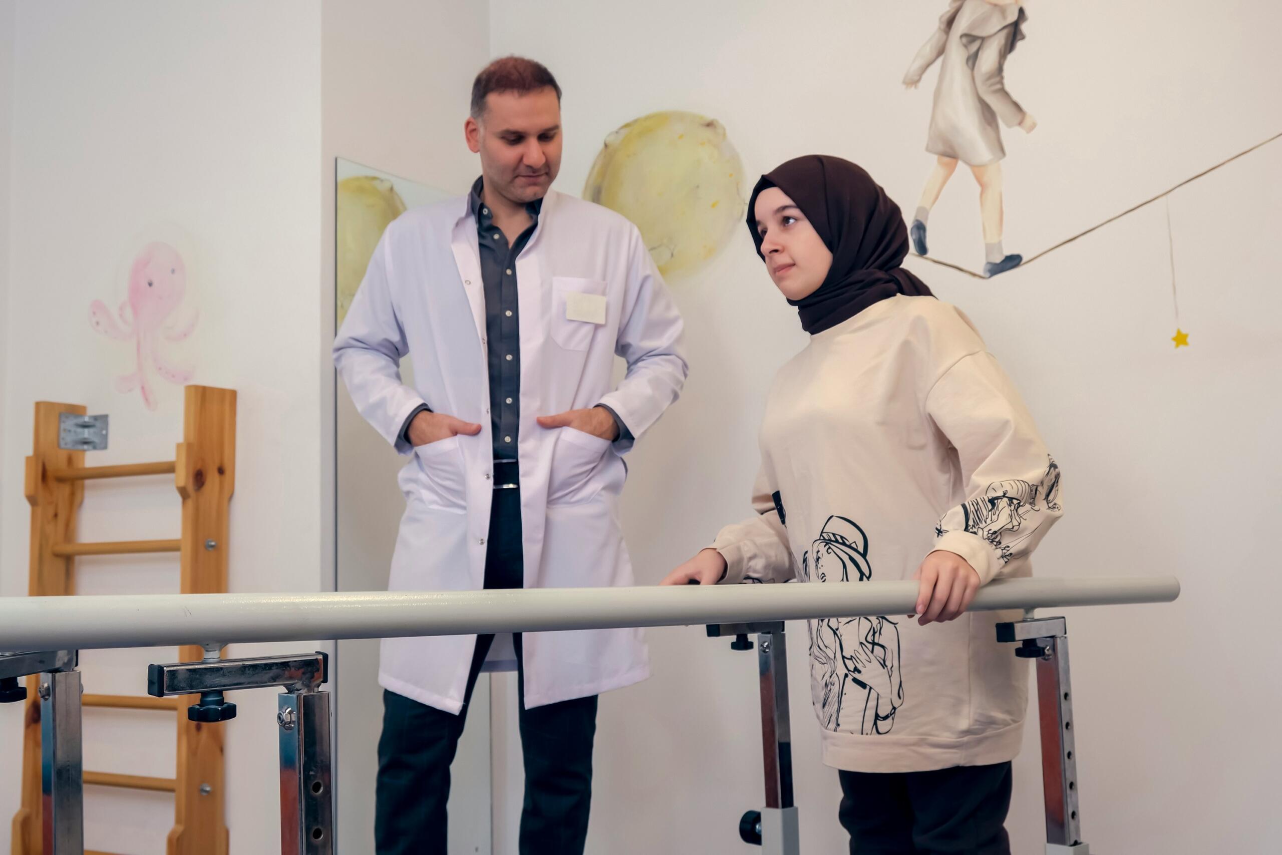 Therapist in white coat observes woman in hijab practicing walking with parallel bars in a decorated therapy room.
