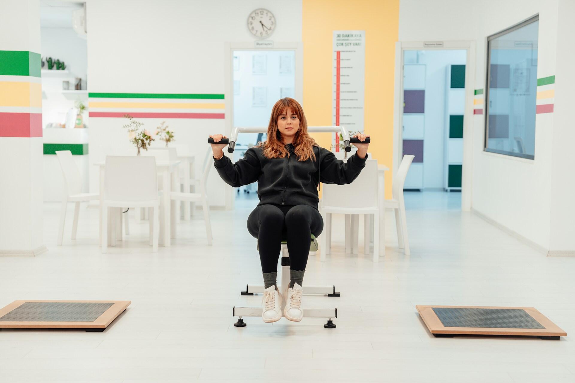 Woman exercising on a seated chest press machine in a bright, colorful physical therapy or fitness room.