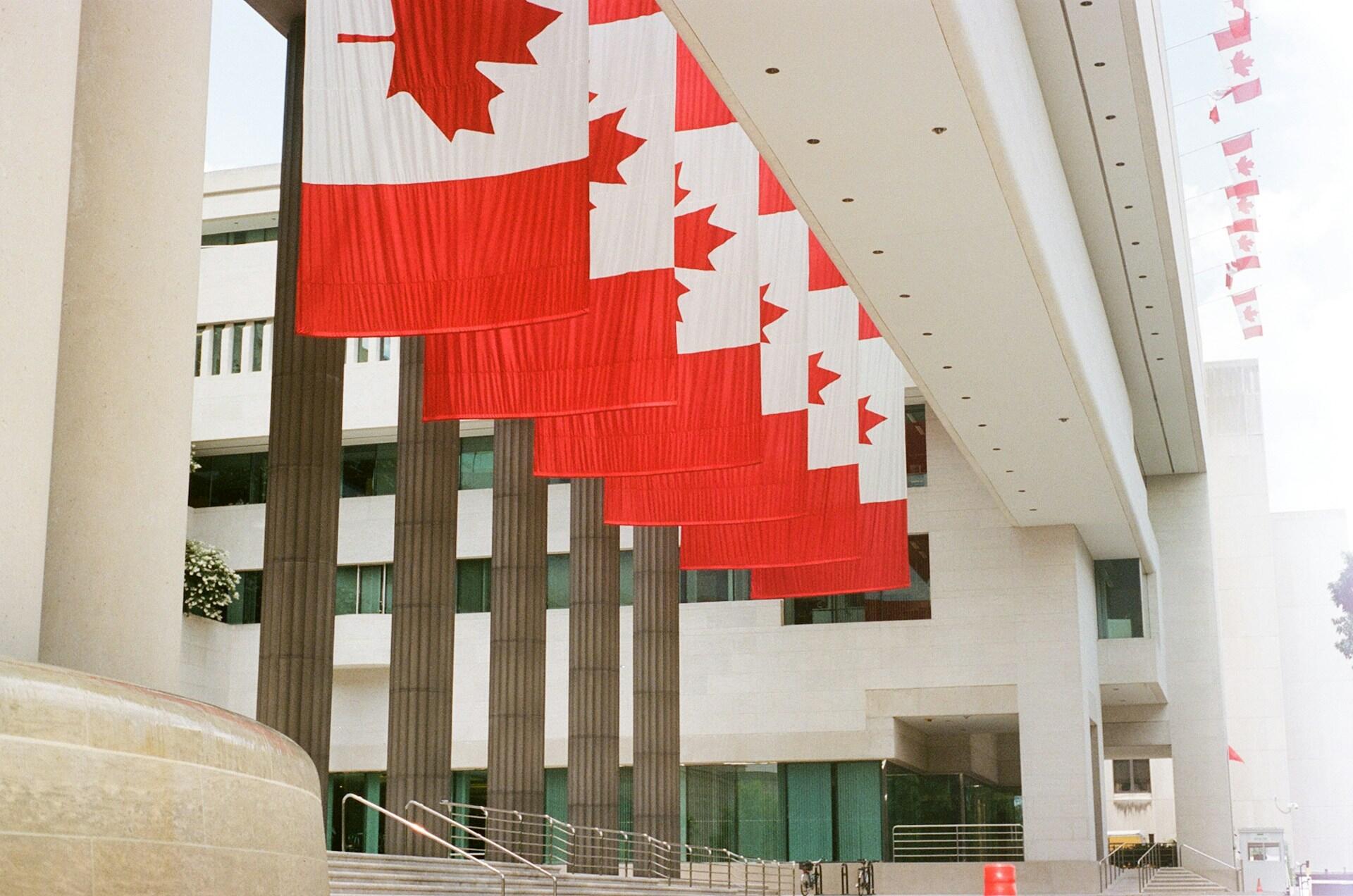 Row of large Canadian flags hanging from a modern building with tall columns and glass windows on a cloudy day.