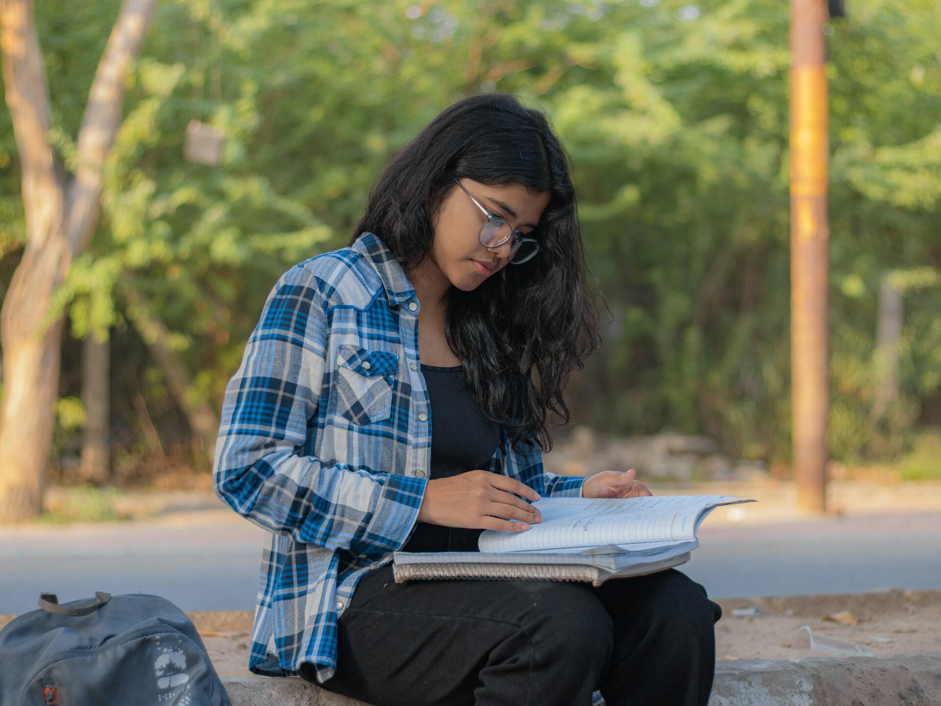 Young woman with glasses reading notes outdoors, wearing a blue plaid shirt with trees and a backpack nearby.