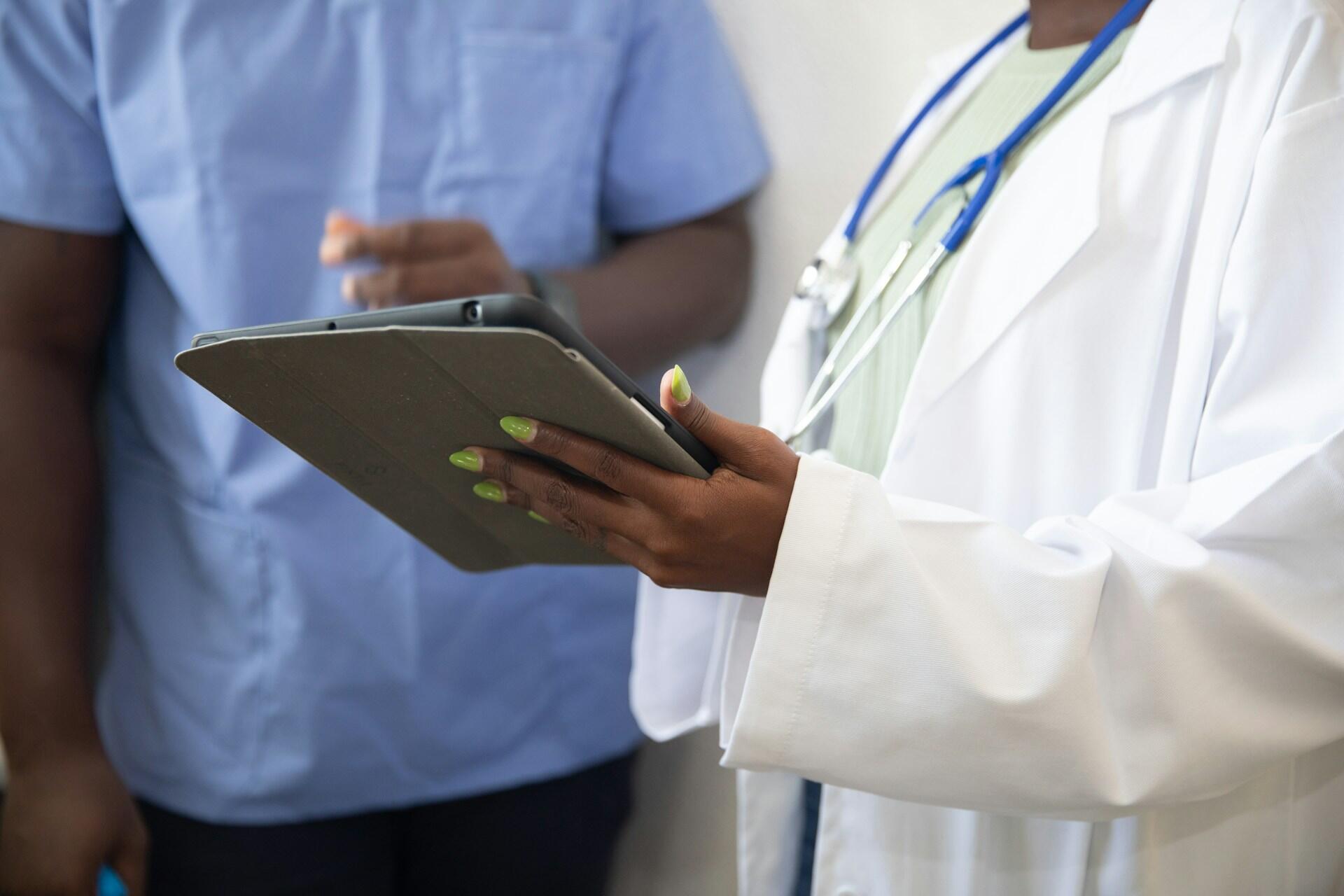 Two medical professionals, one in a white coat with a stethoscope, viewing information on a tablet device.