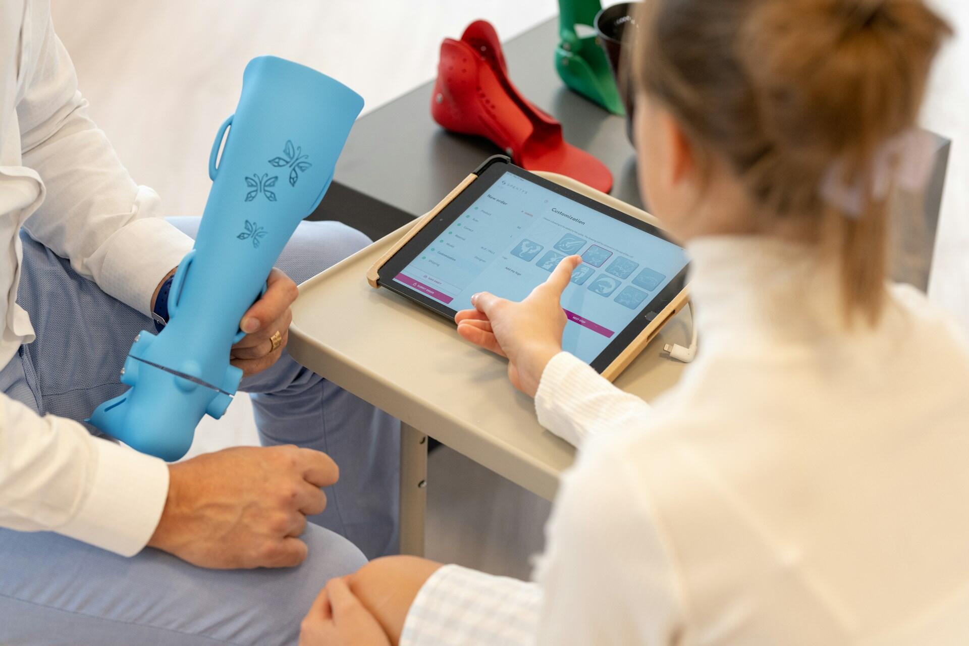 Person customizing a blue orthopedic leg brace with butterfly decals on a tablet, while sitting at a table with red and green braces.