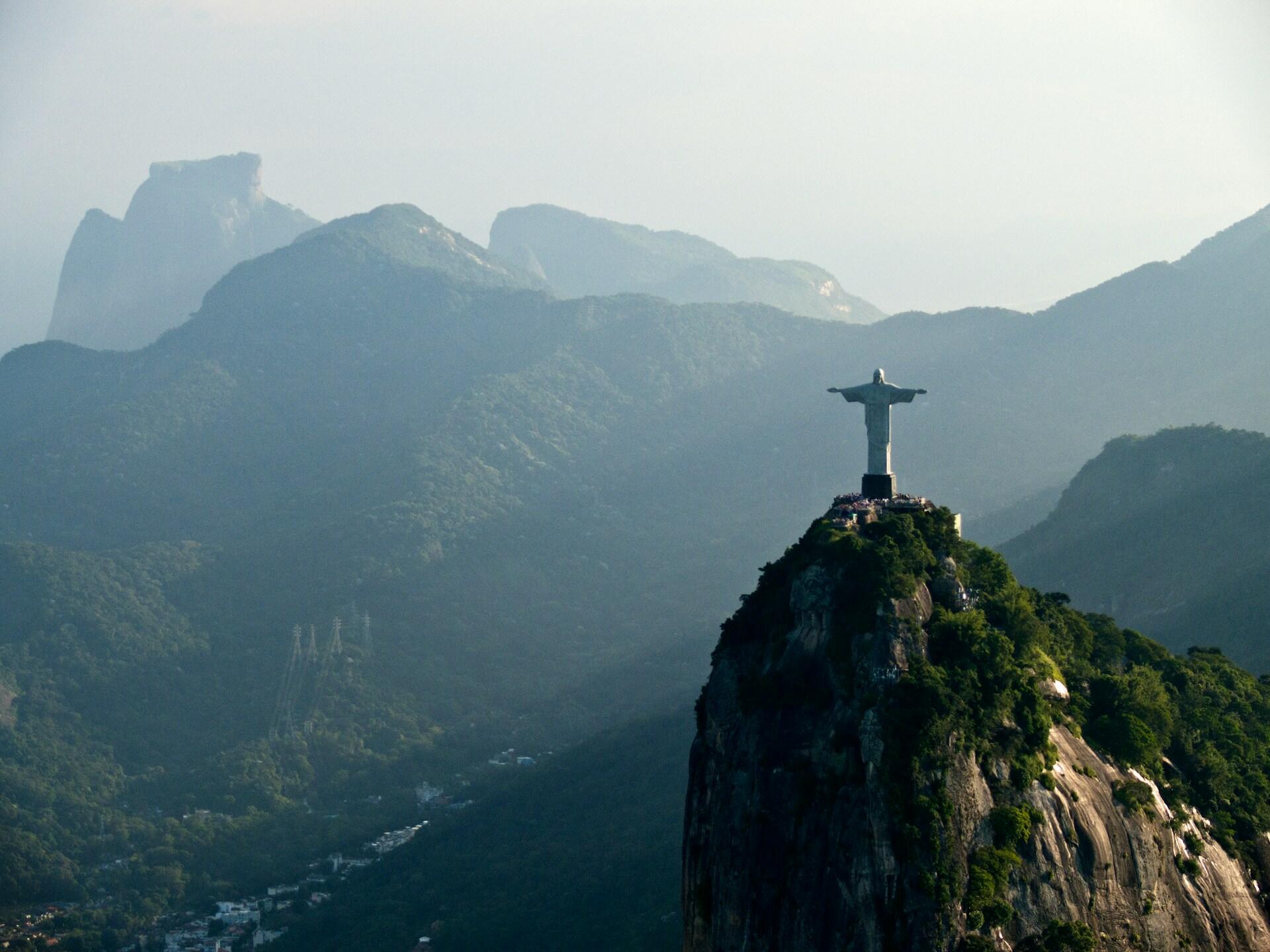 Aerial view of the Christ the Redeemer statue atop a rocky, forested mountain with misty green hills in the background.