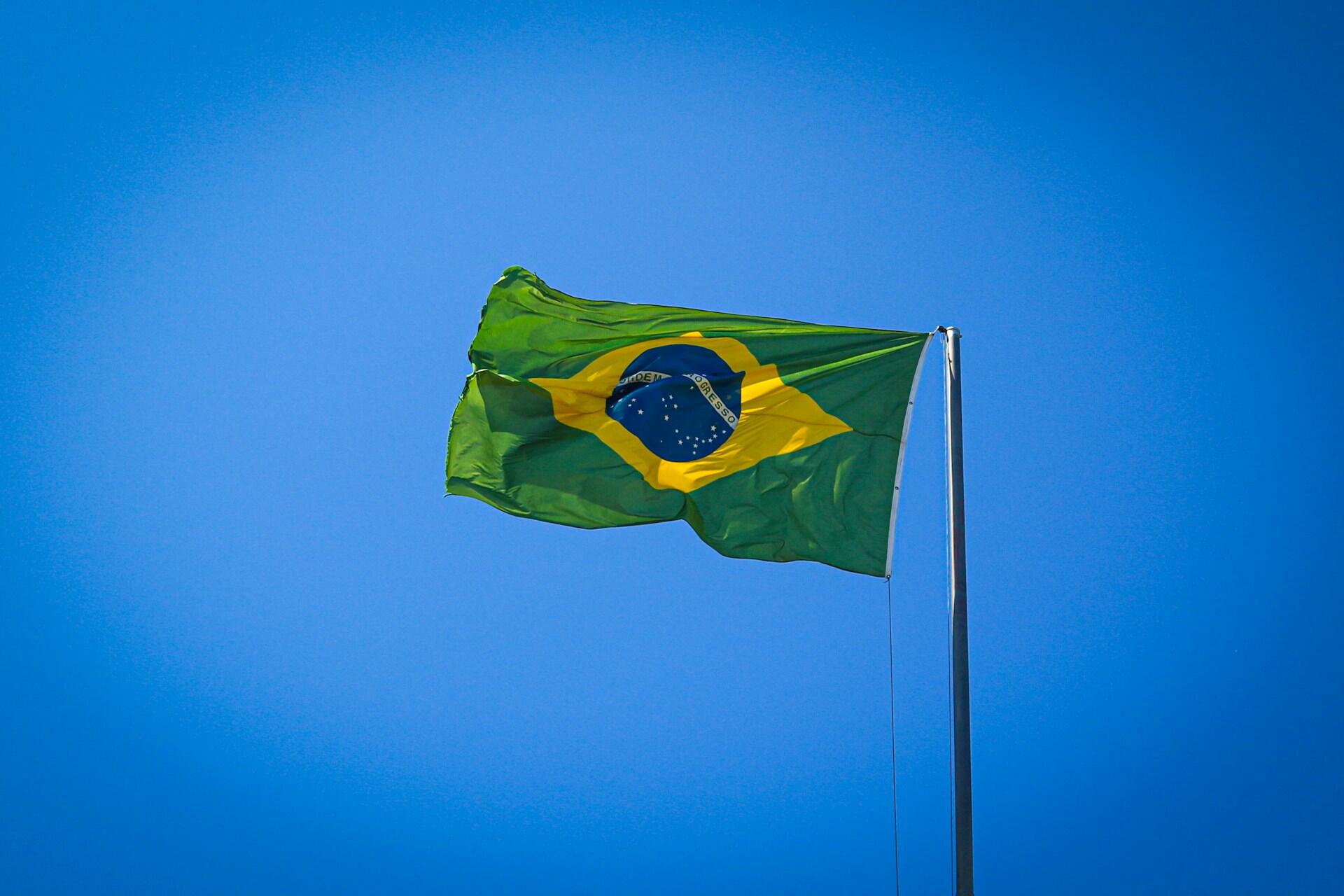 Brazilian flag waving on a tall flagpole against a clear bright blue sky.