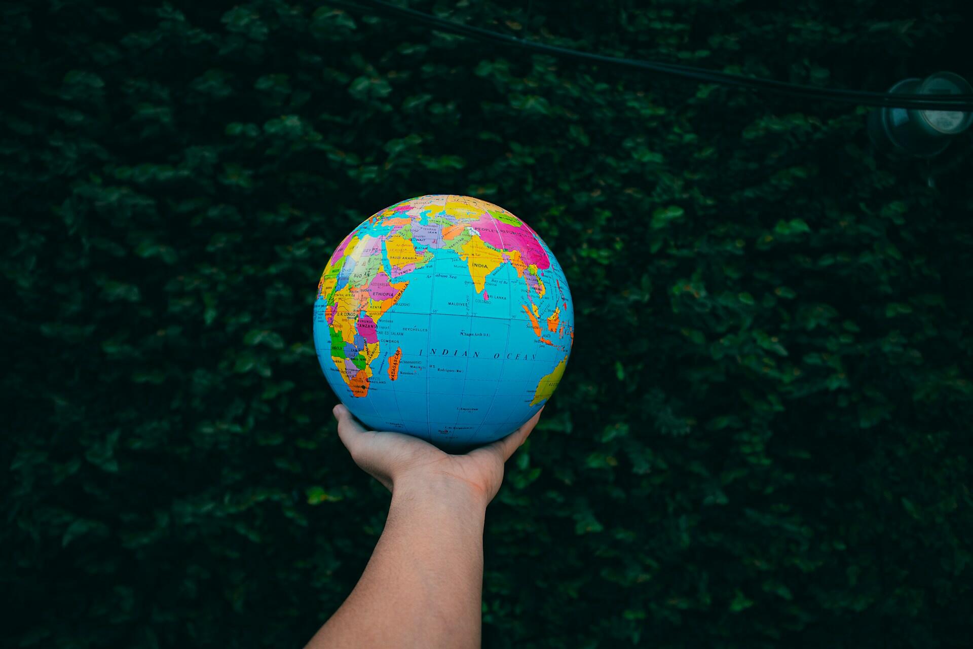 Hand holding a colorful globe showing the Indian Ocean and surrounding continents against a dark leafy background.