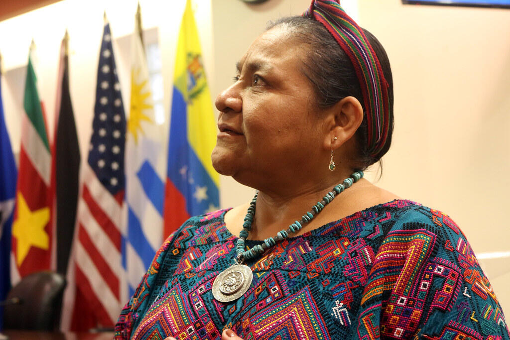 Rigoberta Menchú, Guatemalan Indigenous rights activist and Nobel Peace Prize laureate, wearing traditional Maya clothing and turquoise jewelry while speaking at an event. Flags of several countries are displayed in the background.