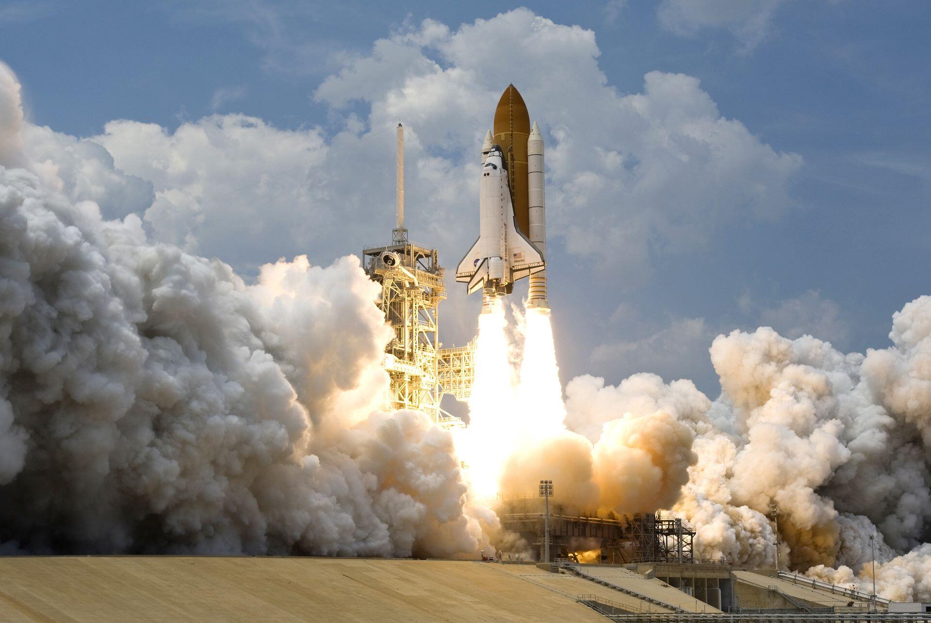 NASA's Space Shuttle launches from the launch pad, with bright rocket flames and thick clouds of smoke billowing beneath it as it ascends into a partly cloudy sky.