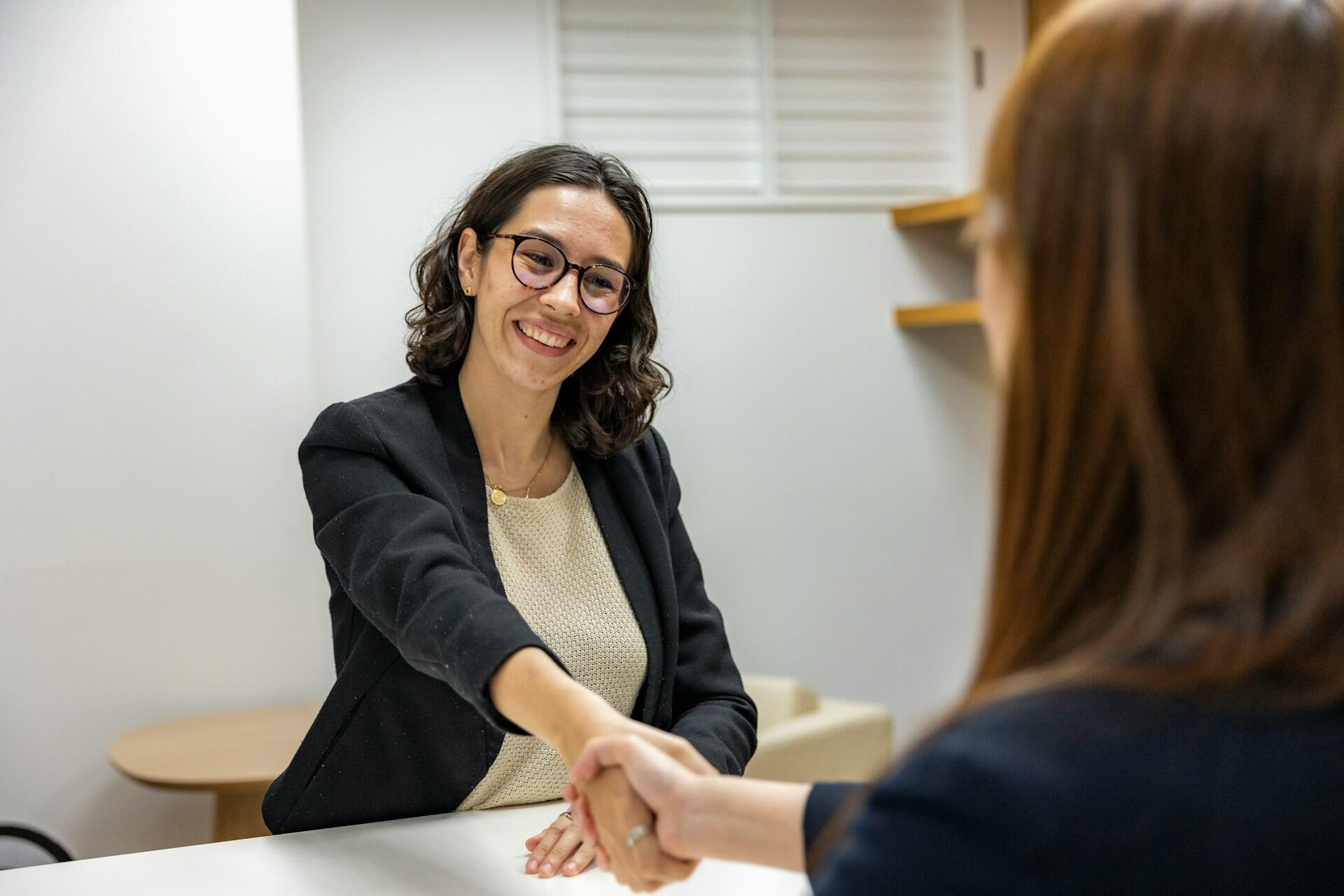 Two women shaking hands across a table in a bright office, one smiling and wearing glasses and a black blazer.