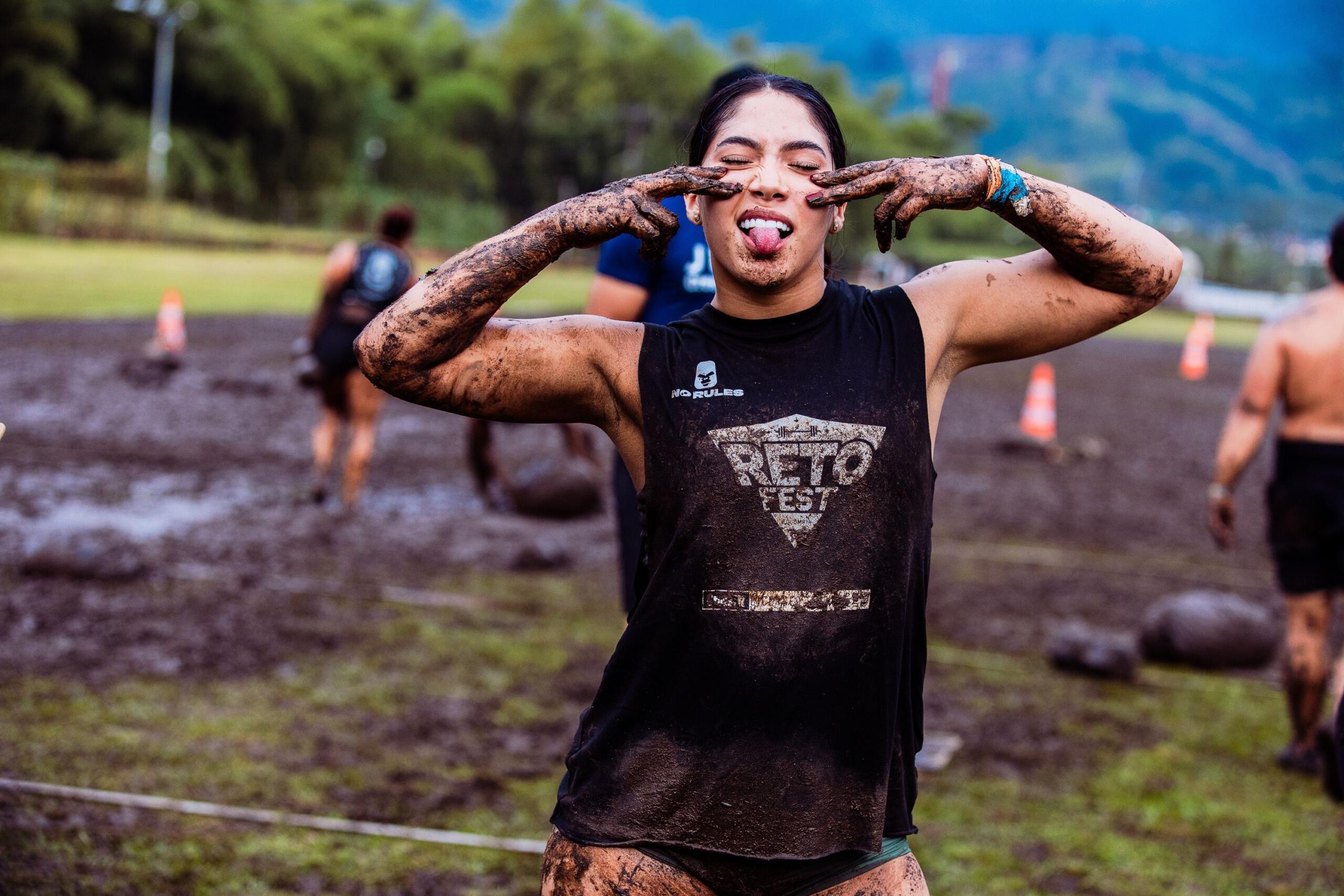 An obstacle course athlete covered in mud sticks her tongue out playfully, enjoying the moment. She wears a black sleeveless top and stands in a field of mud.