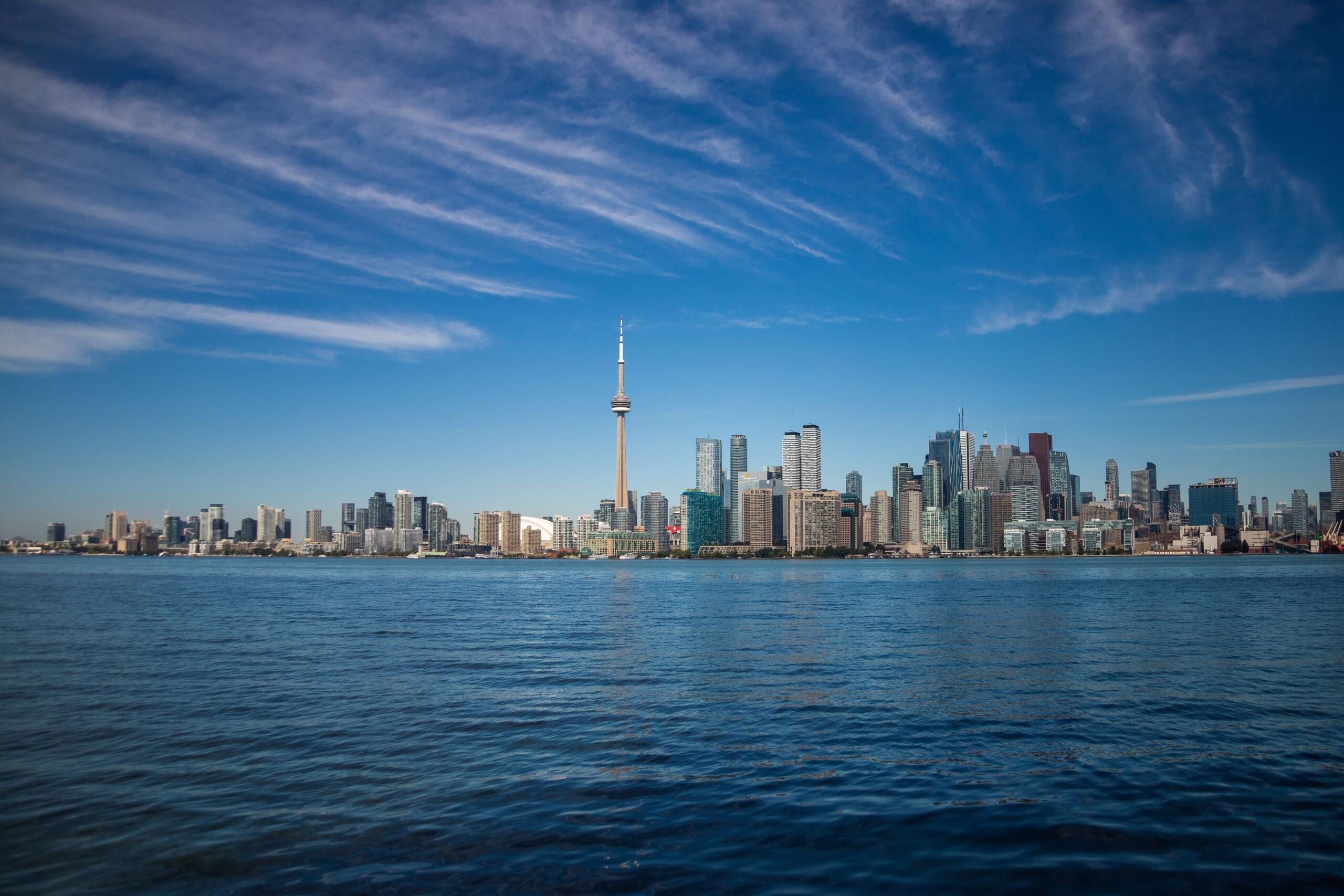 A clear daytime view of Toronto's skyline under blue skies, with the CN Tower rising above modern skyscrapers and Lake Ontario in the foreground on a bright, sunny day.