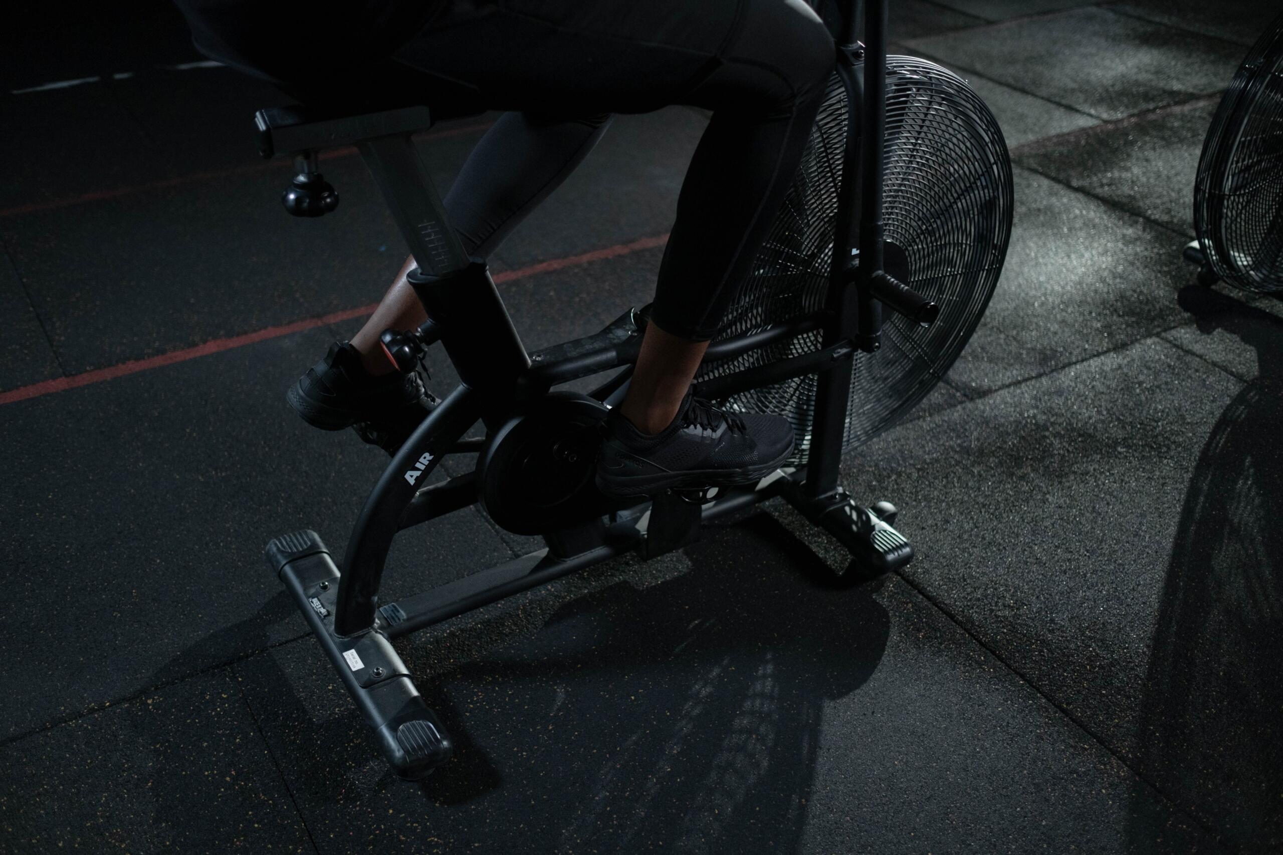 A man rides an air bike in a dimly lit gym, leaning forward as he powers through an intense cardio workout.