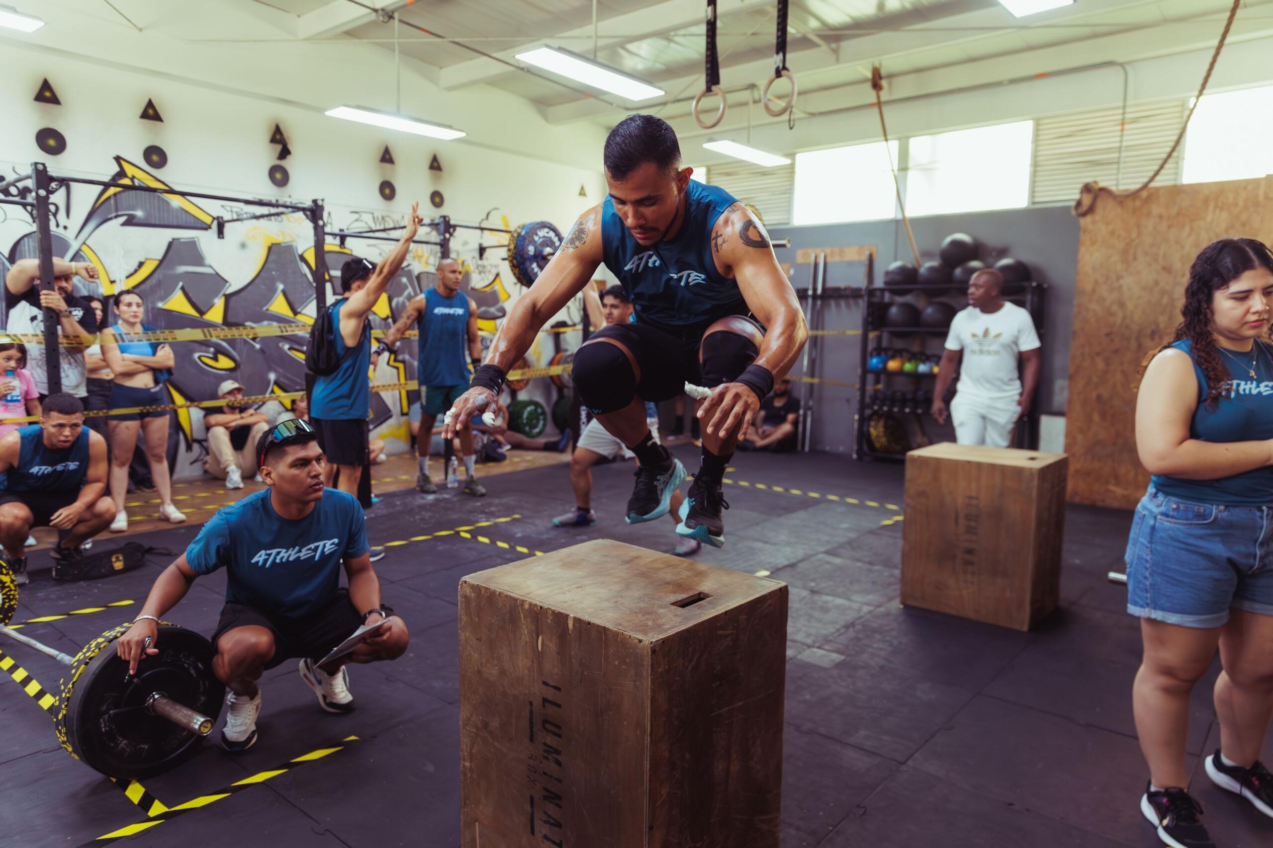 A man performs box jumps in a busy gym, explosively jumping onto a plyometric box while other people train on fitness equipment in the background.