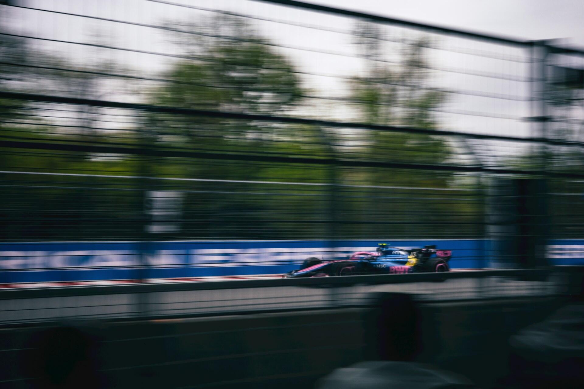 Blurred photo of a Formula 1 race car speeding past a fenced track with spectators in the foreground.