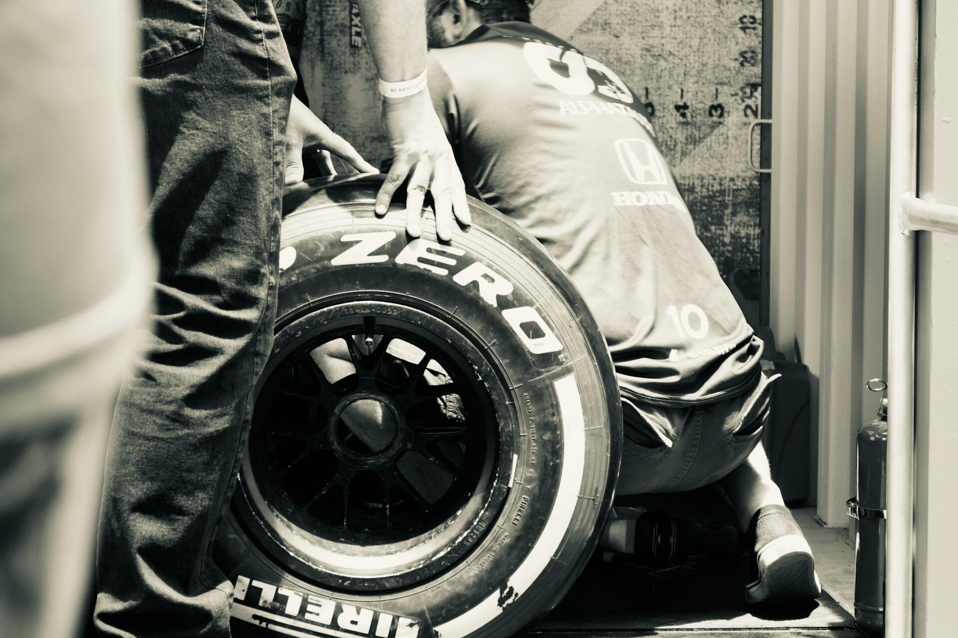 Two mechanics changing or inspecting a Pirelli racing tire in a garage or pit area.