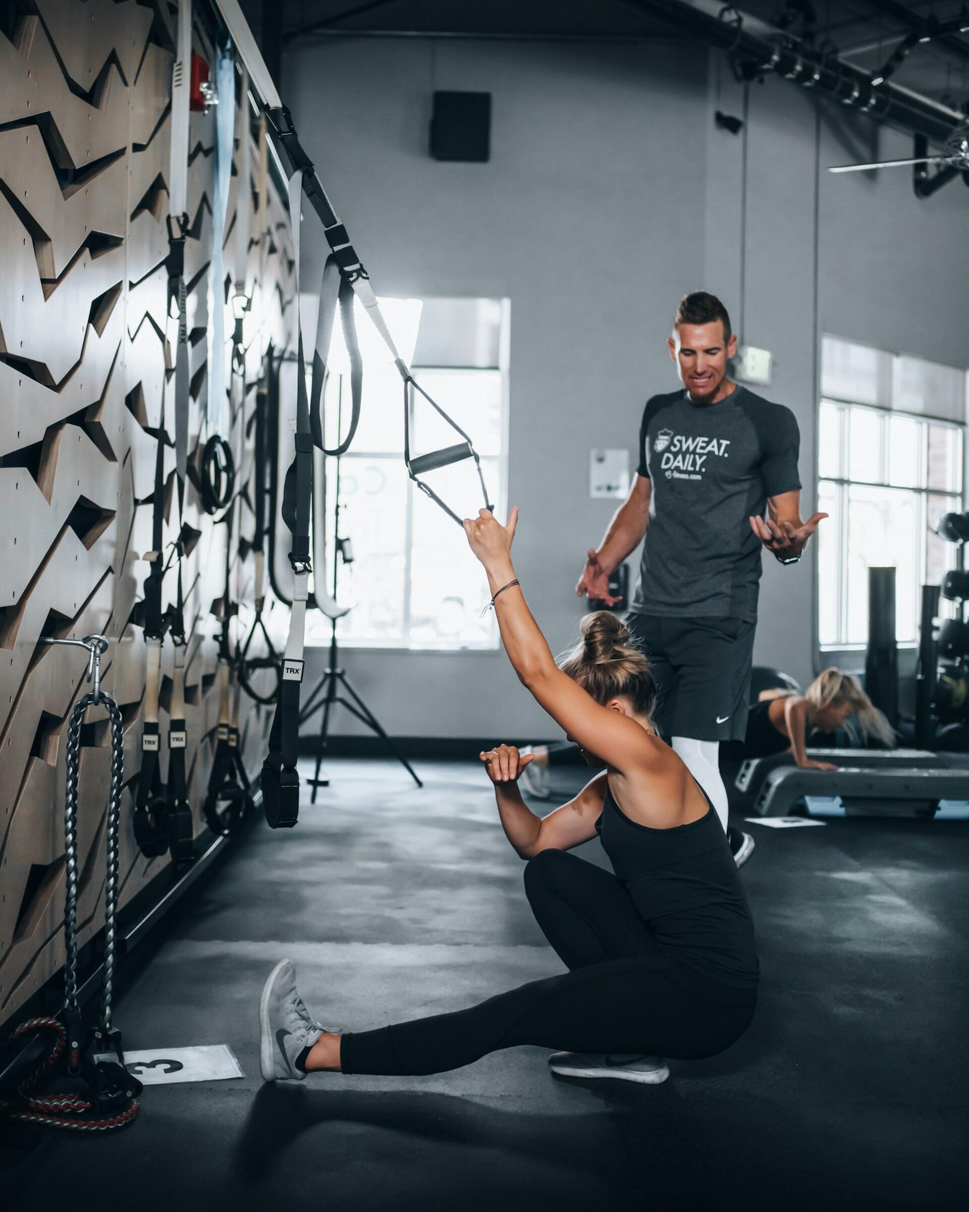 Woman using TRX straps for a one-legged squat exercise while a male trainer observes and instructs in a gym setting.