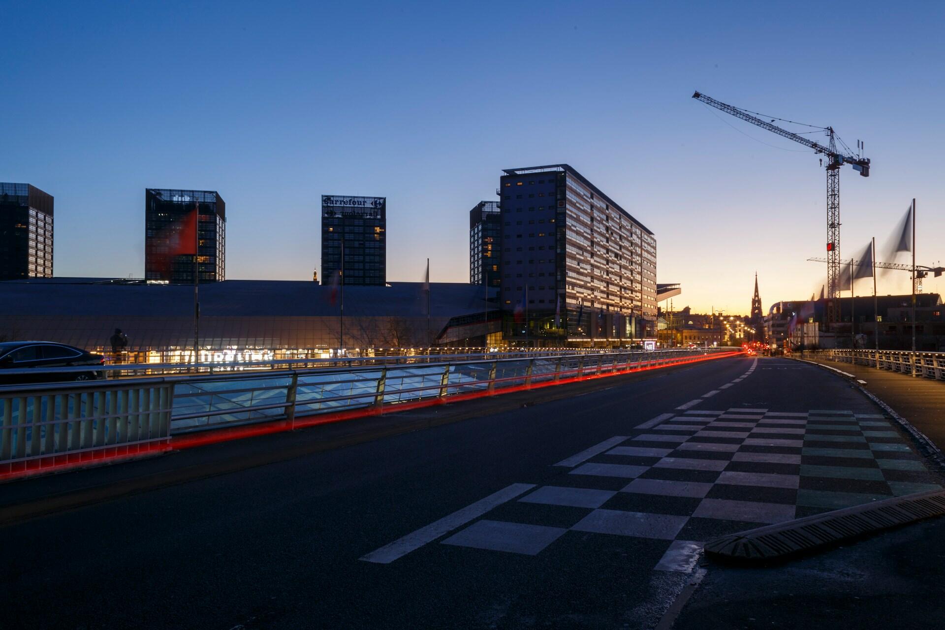 Urban street at dusk with modern buildings, a construction crane, light trails from vehicles, and a church spire in the background.