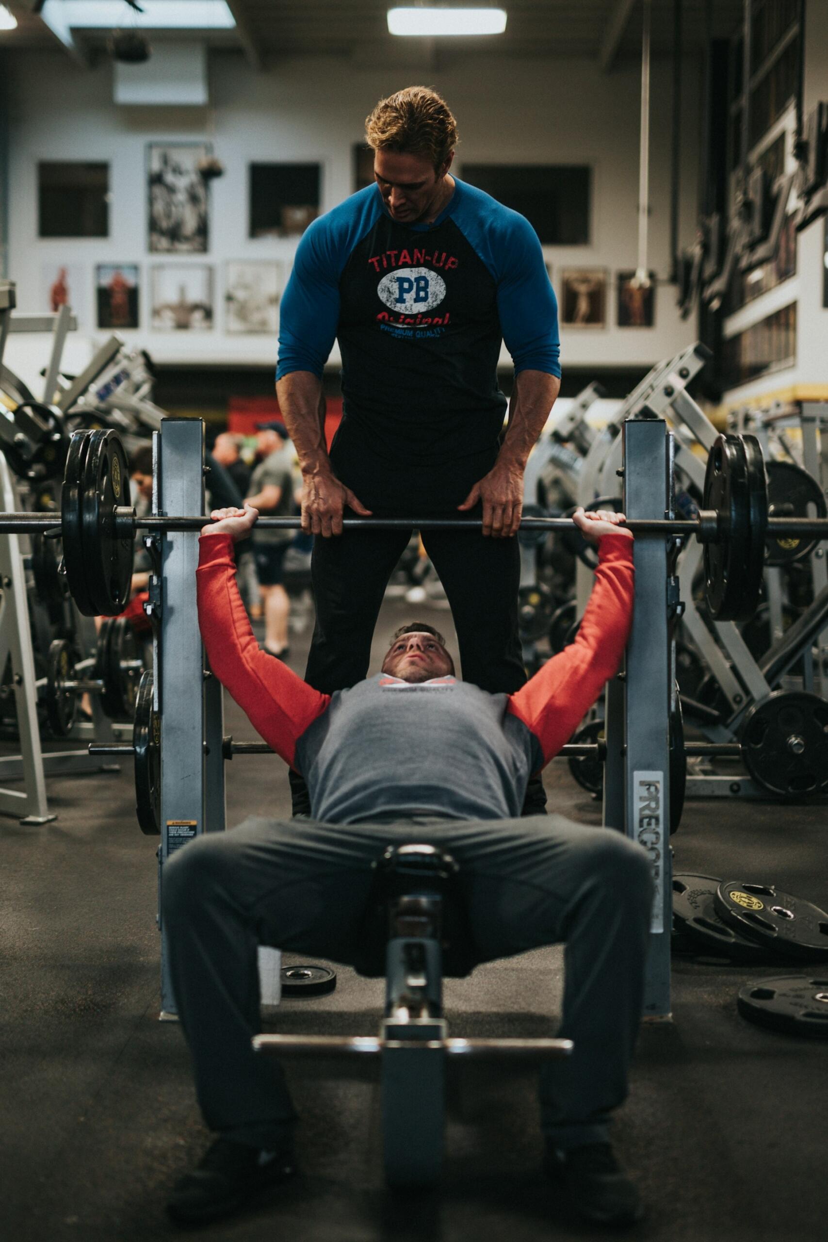 Man spotting another man lifting a barbell on a bench press in a busy gym filled with weightlifting equipment.