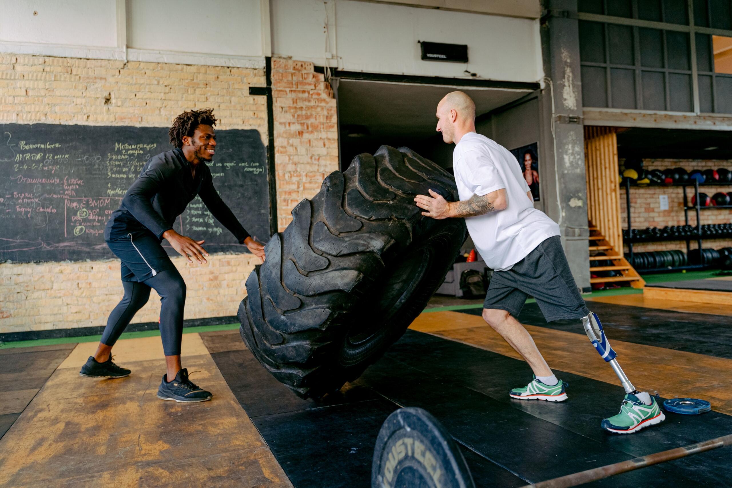 Two men lift and hold a large training tire upright in a gym, working together during a functional fitness strength exercise.