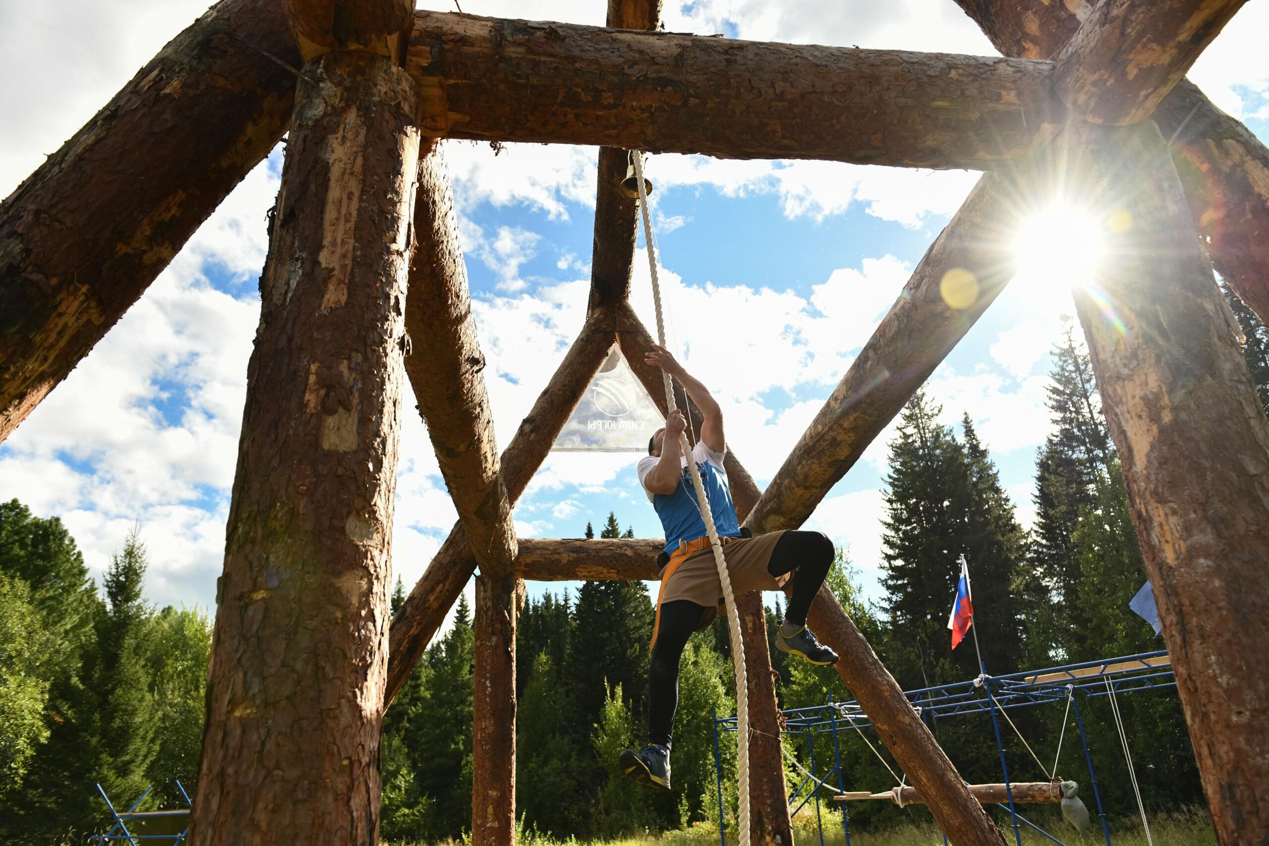 A man hangs from a rope beneath a large wooden obstacle, using his upper-body strength to swing across during an outdoor obstacle course race.