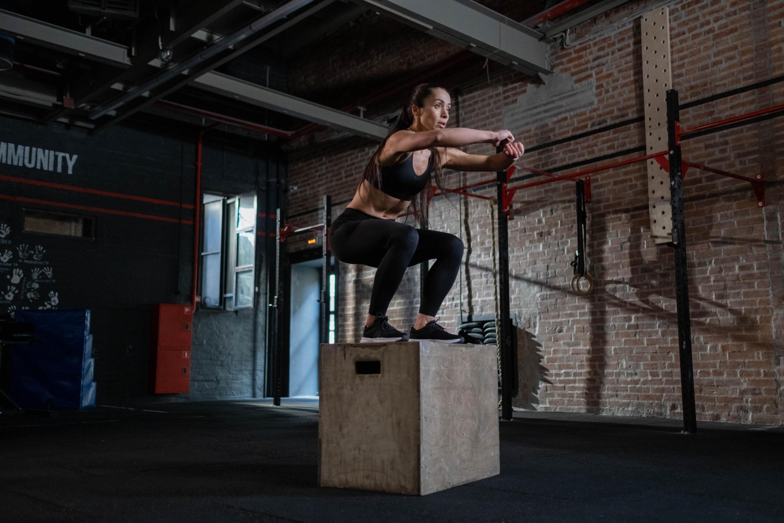 A woman performs a box jump in a gym, explosively jumping onto a plyometric box with focused form during a strength and conditioning workout.