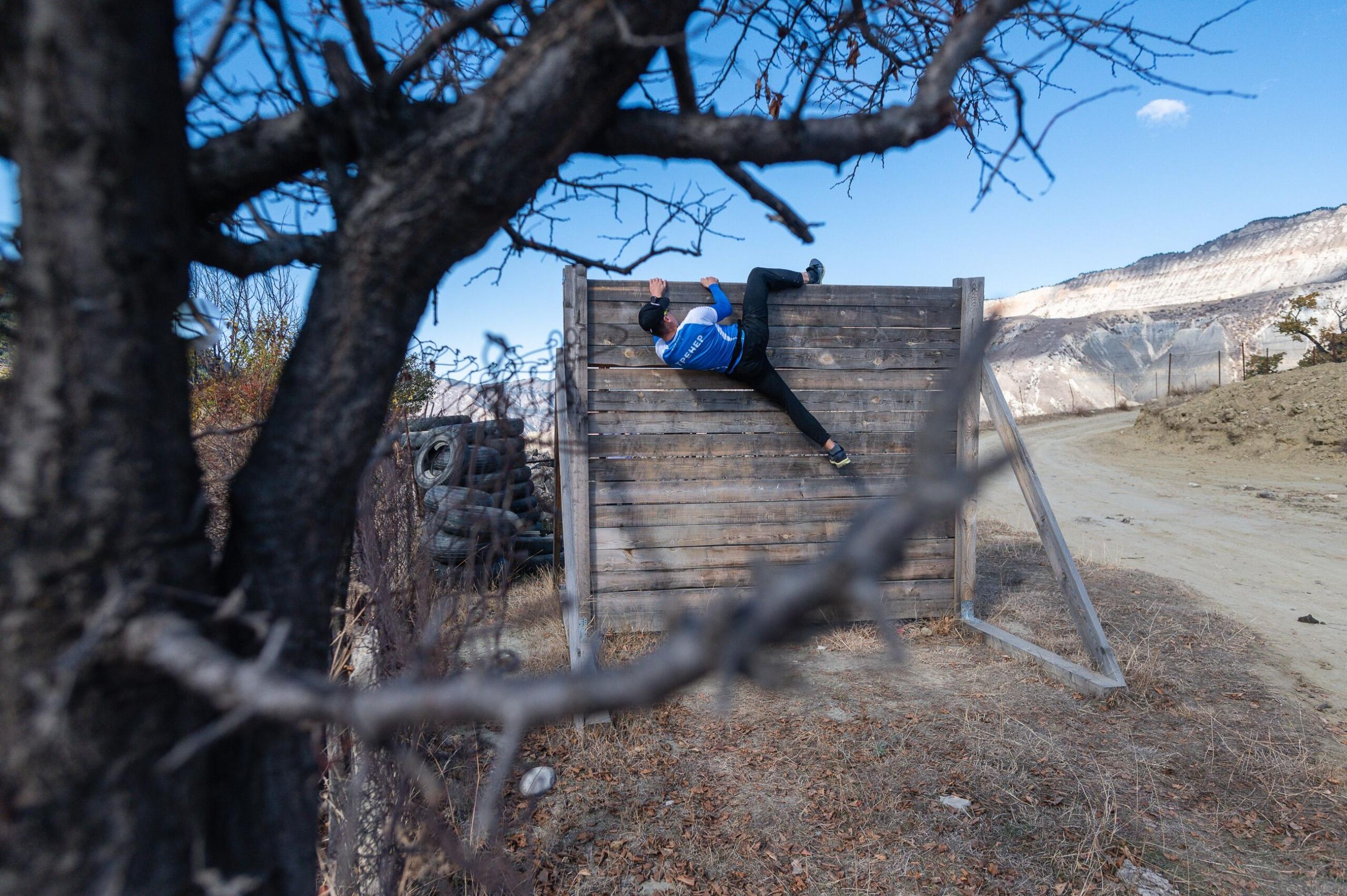 A man climbs over a large wooden obstacle during an obstacle course race, using both hands and feet to pull himself over the structure.