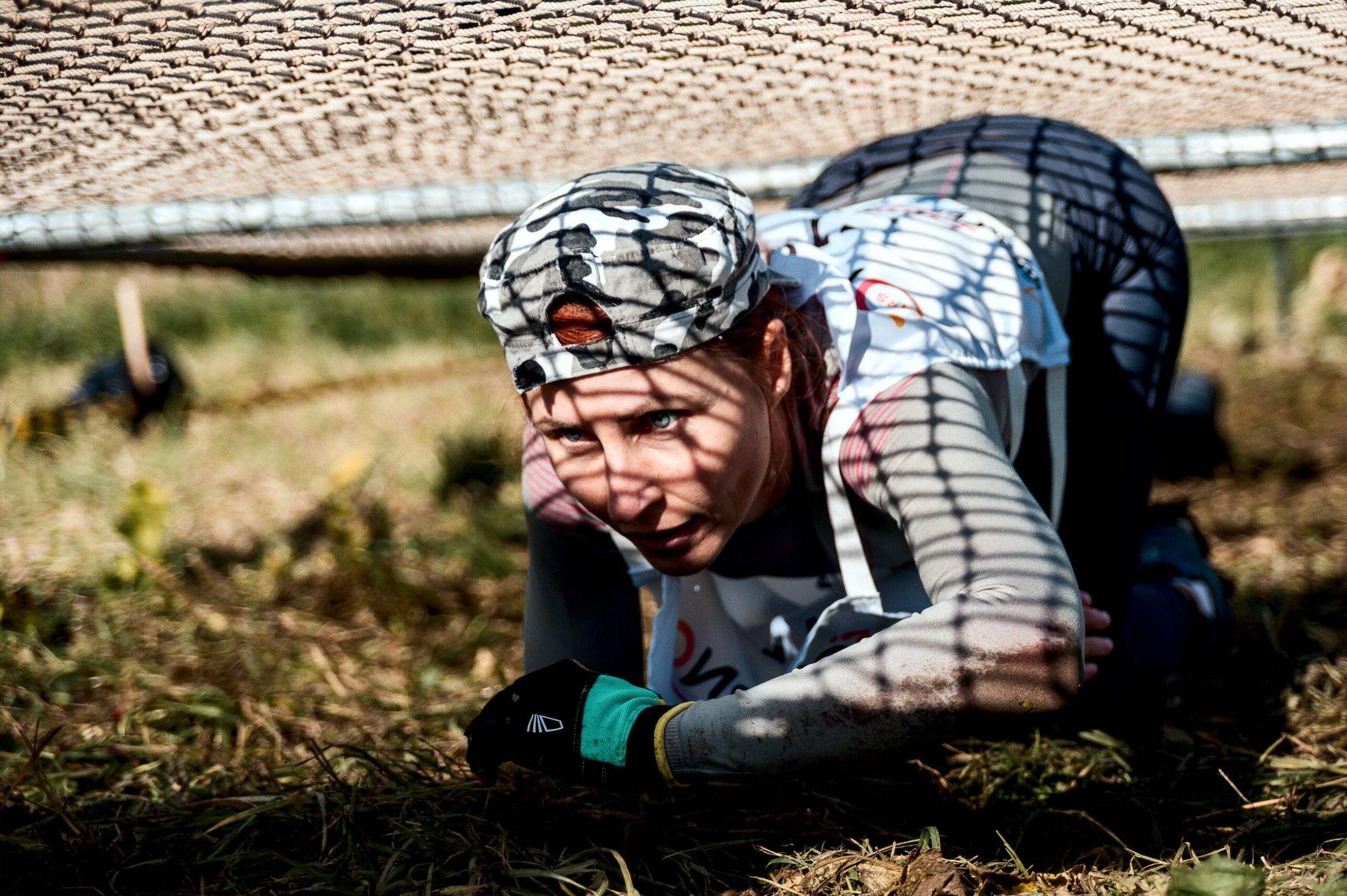 A woman crawls beneath a low obstacle on a muddy obstacle course, concentrating as she navigates the challenge during an outdoor endurance race.