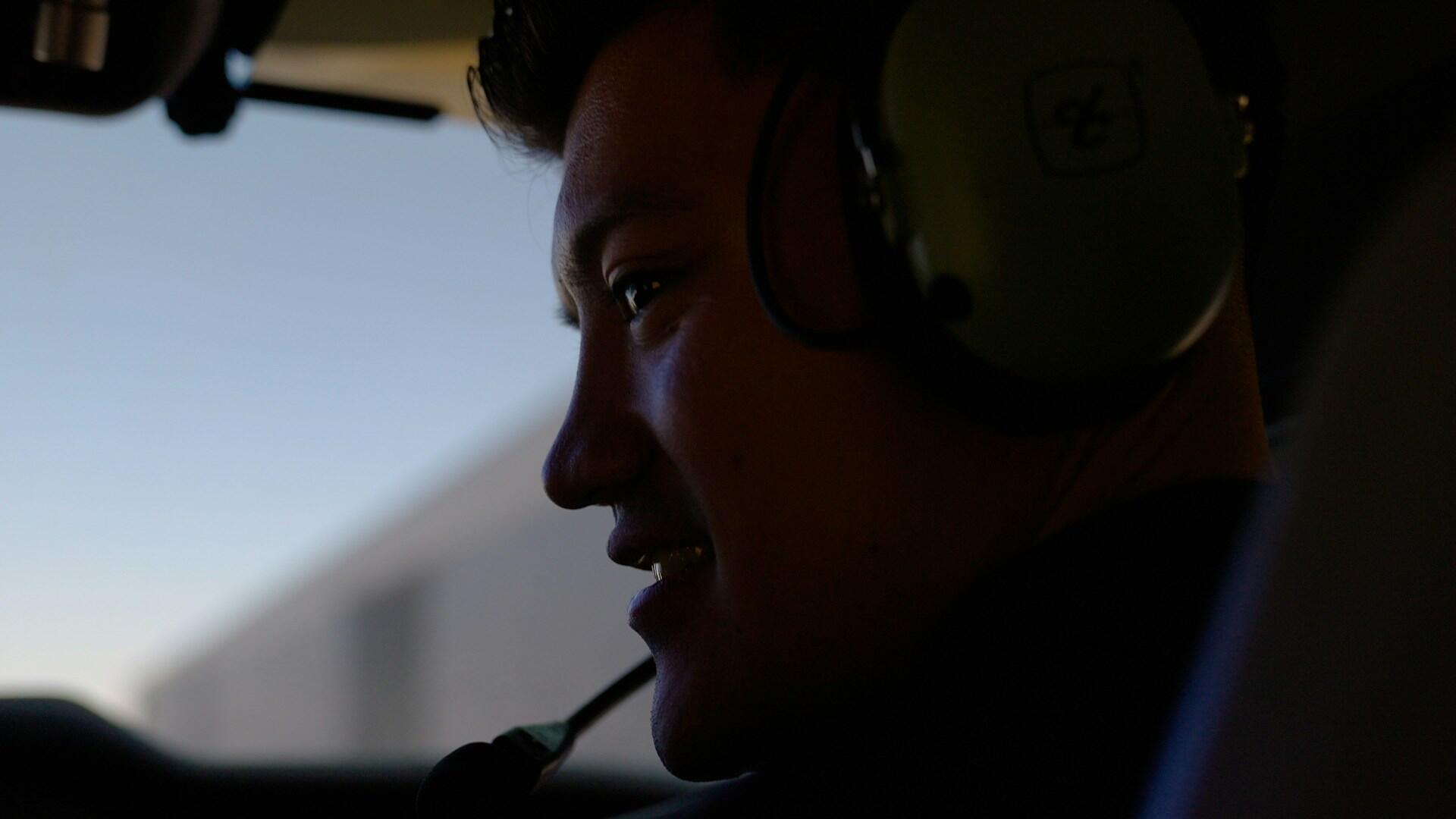 Close-up of a person wearing a pilot headset, smiling, inside a cockpit with a window showing a blurred outdoor scene.