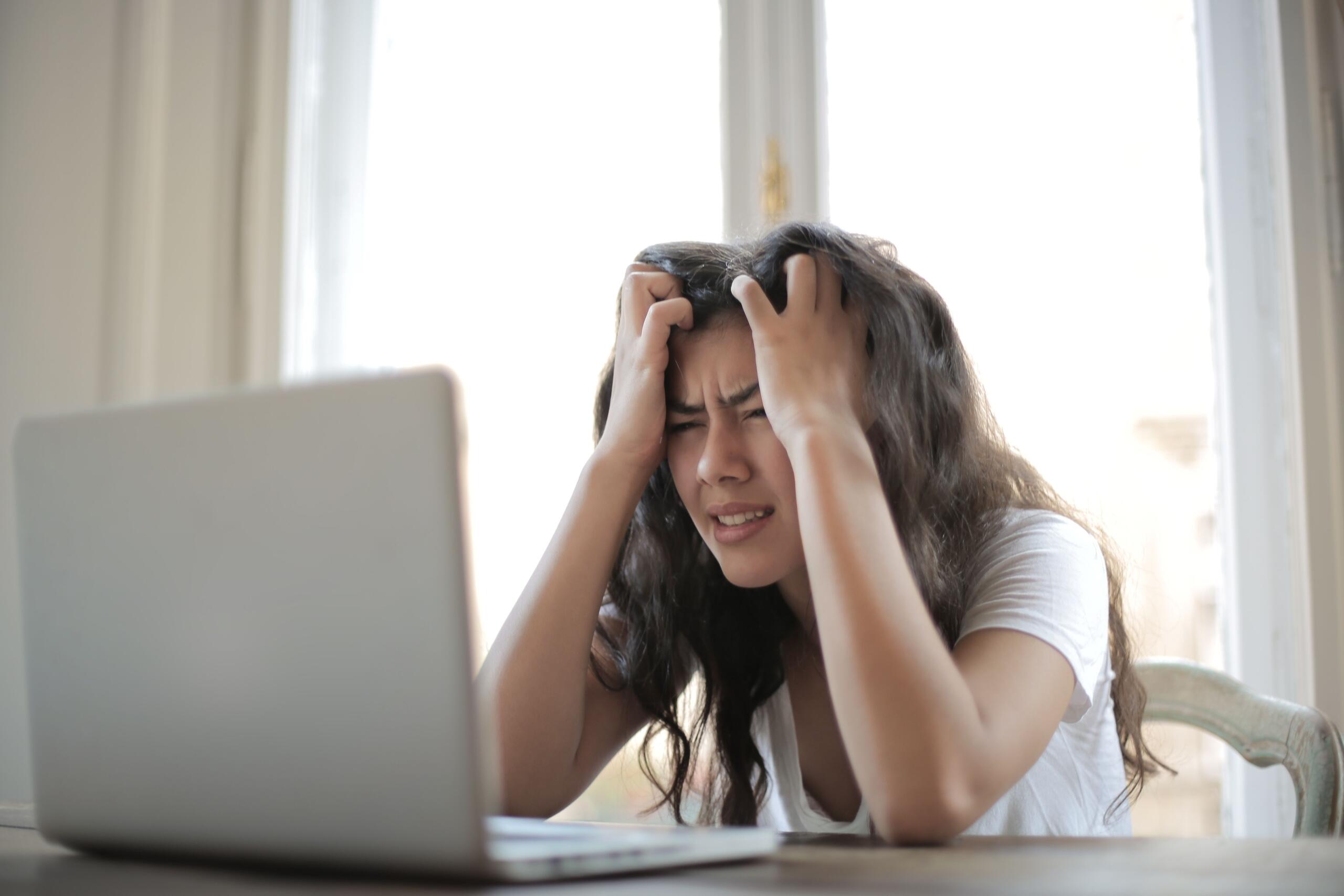 A young woman holds her head in her hands, appearing stressed as she looks at her computer.