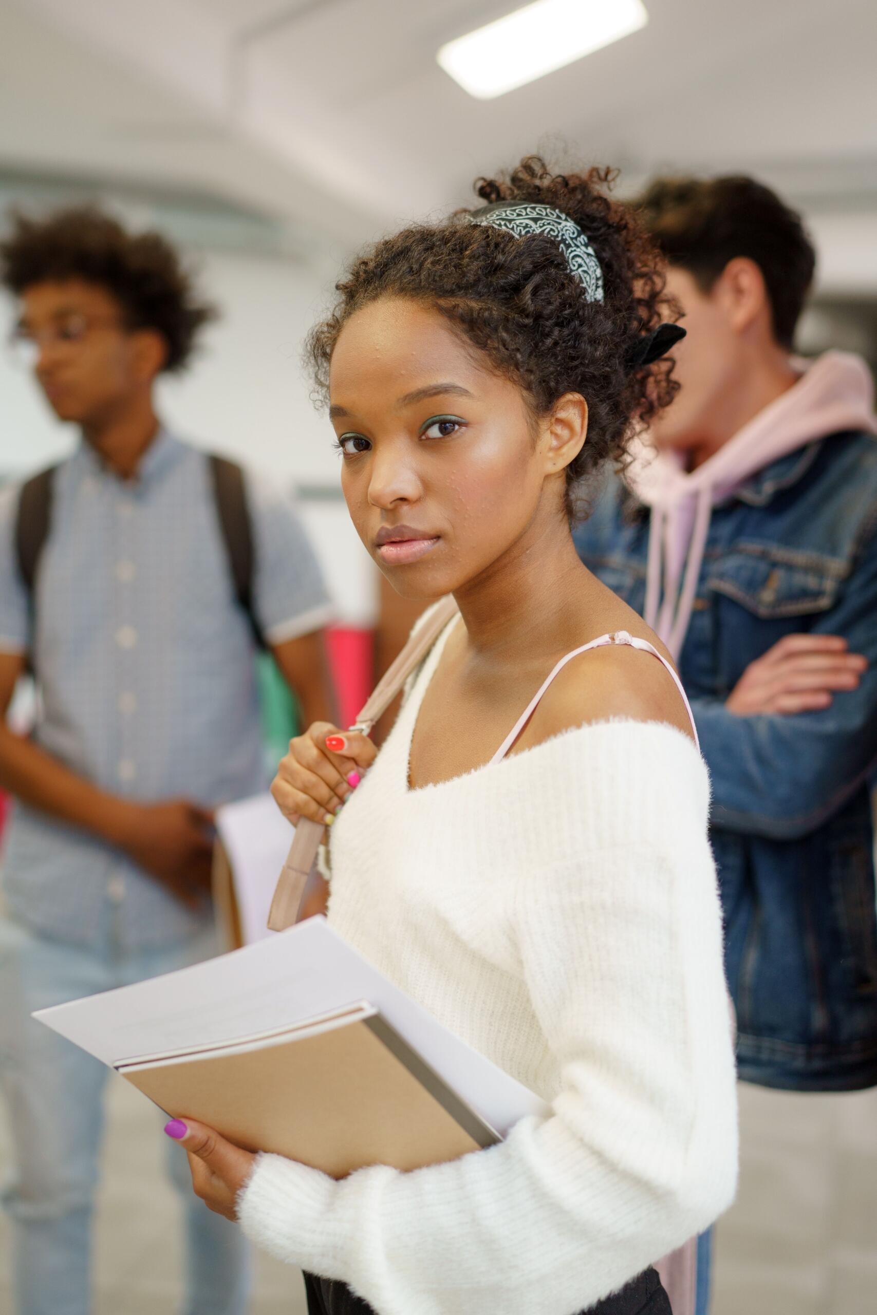 A young woman near high school graduation looks ahead, holding her notebooks while surrounded by students.