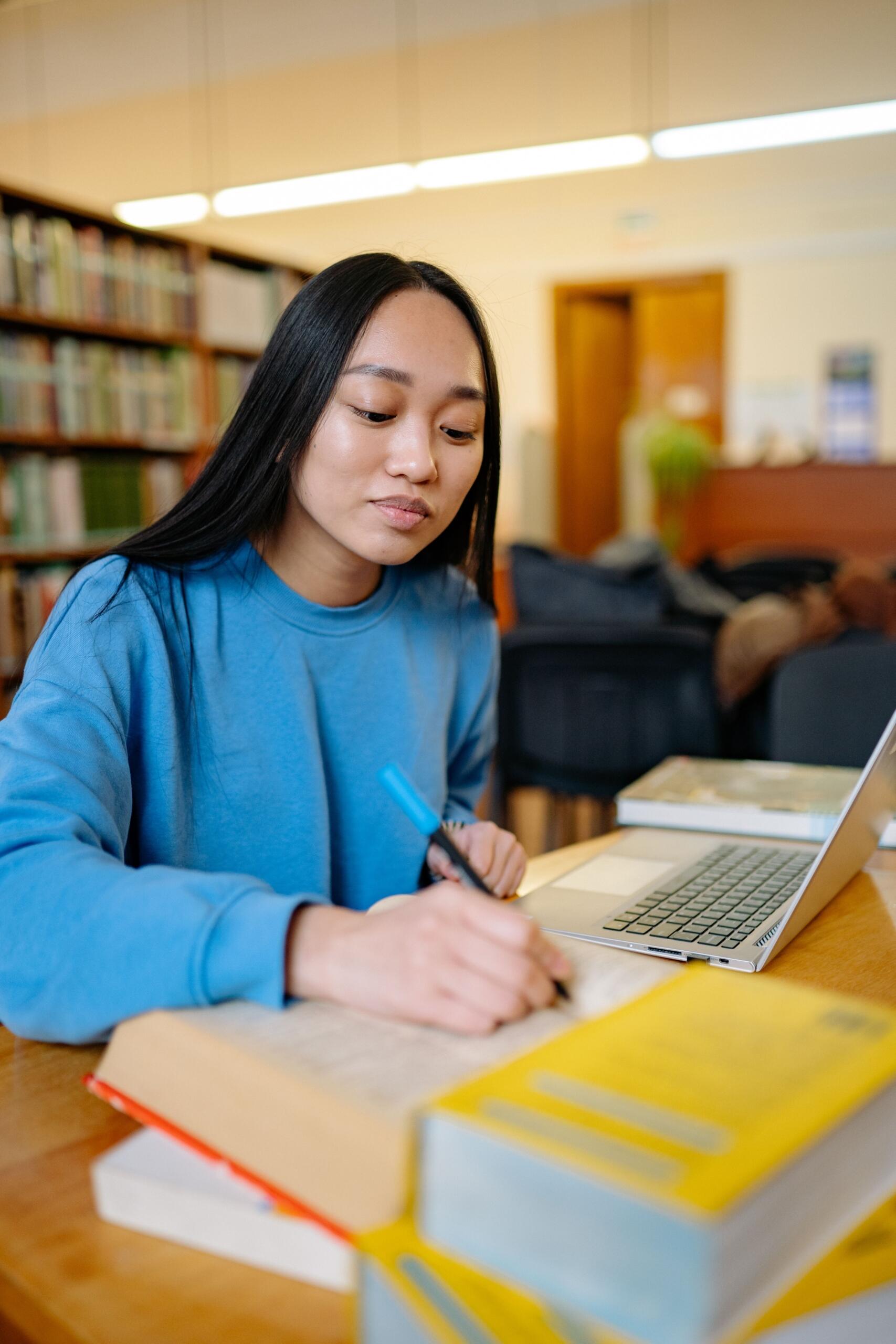 A young adult female working in a library. She is focused as she writes notes and studies a text beside a pile of books.