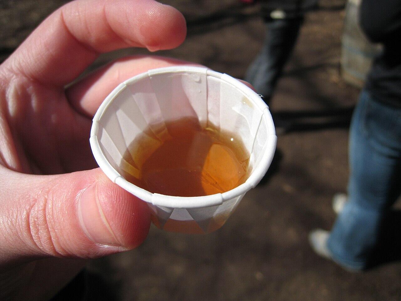 Hand holding a small paper cup filled with amber-colored liquid, with blurred people in the background.