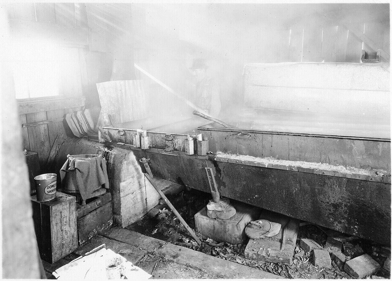 Black and white photo of an old sugarhouse evaporator with steam and a worker stirring inside a wooden building.