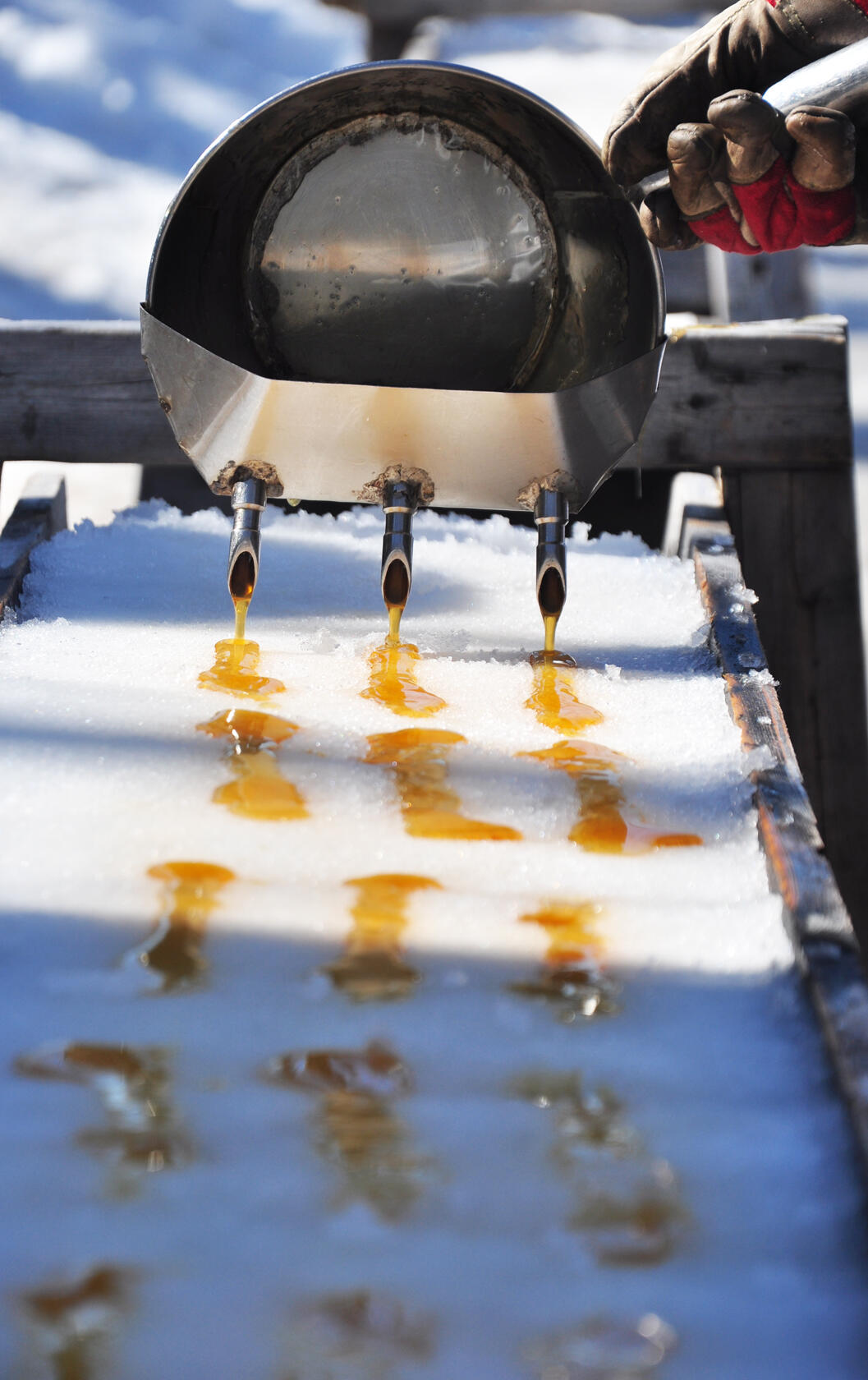 Maple sap syrup is being poured through three spouts onto a snow-covered wooden board outdoors.