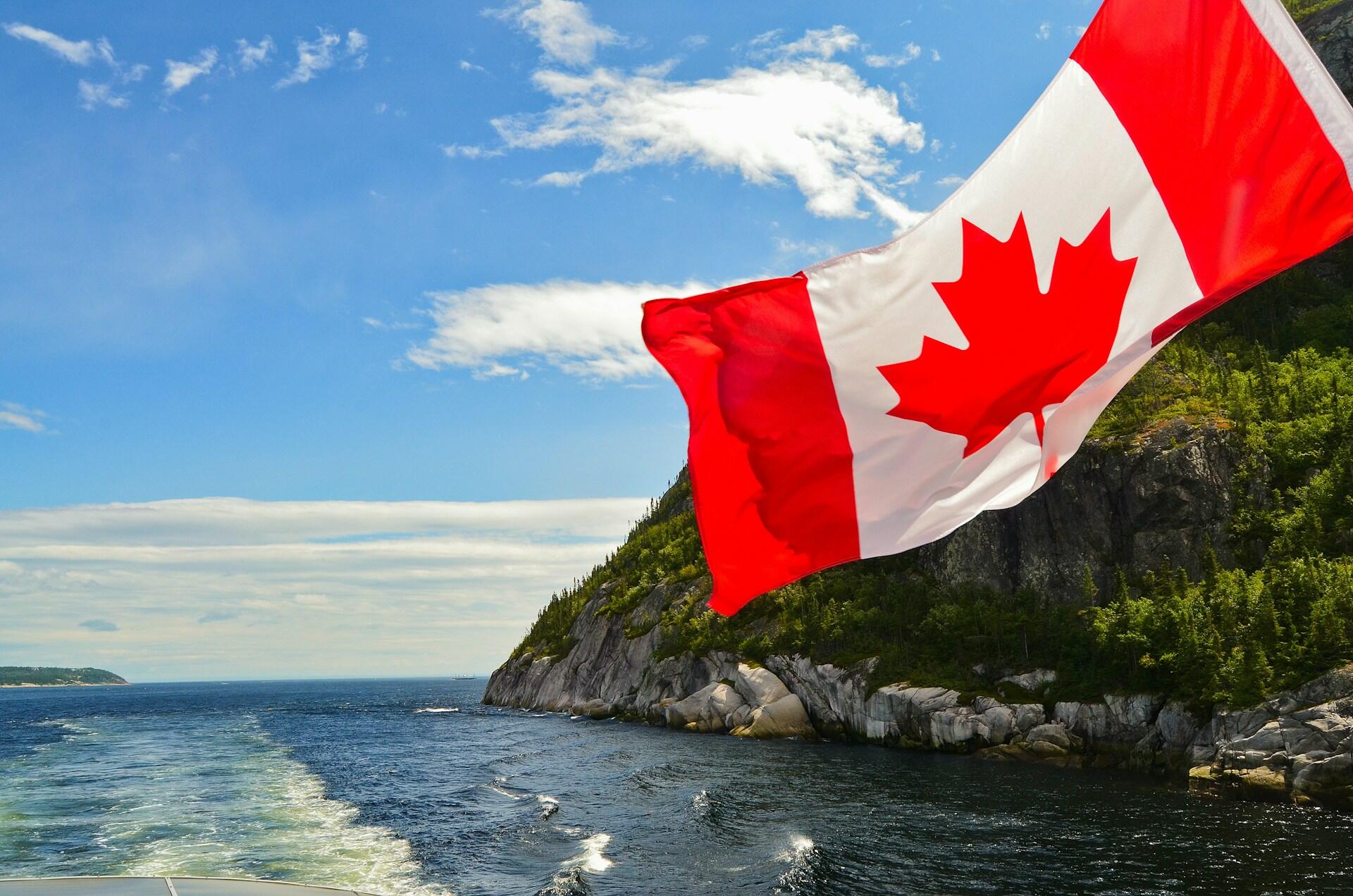 Canadian flag waving in the wind with a picturesque landscape of ocean waves and cliffs in the background