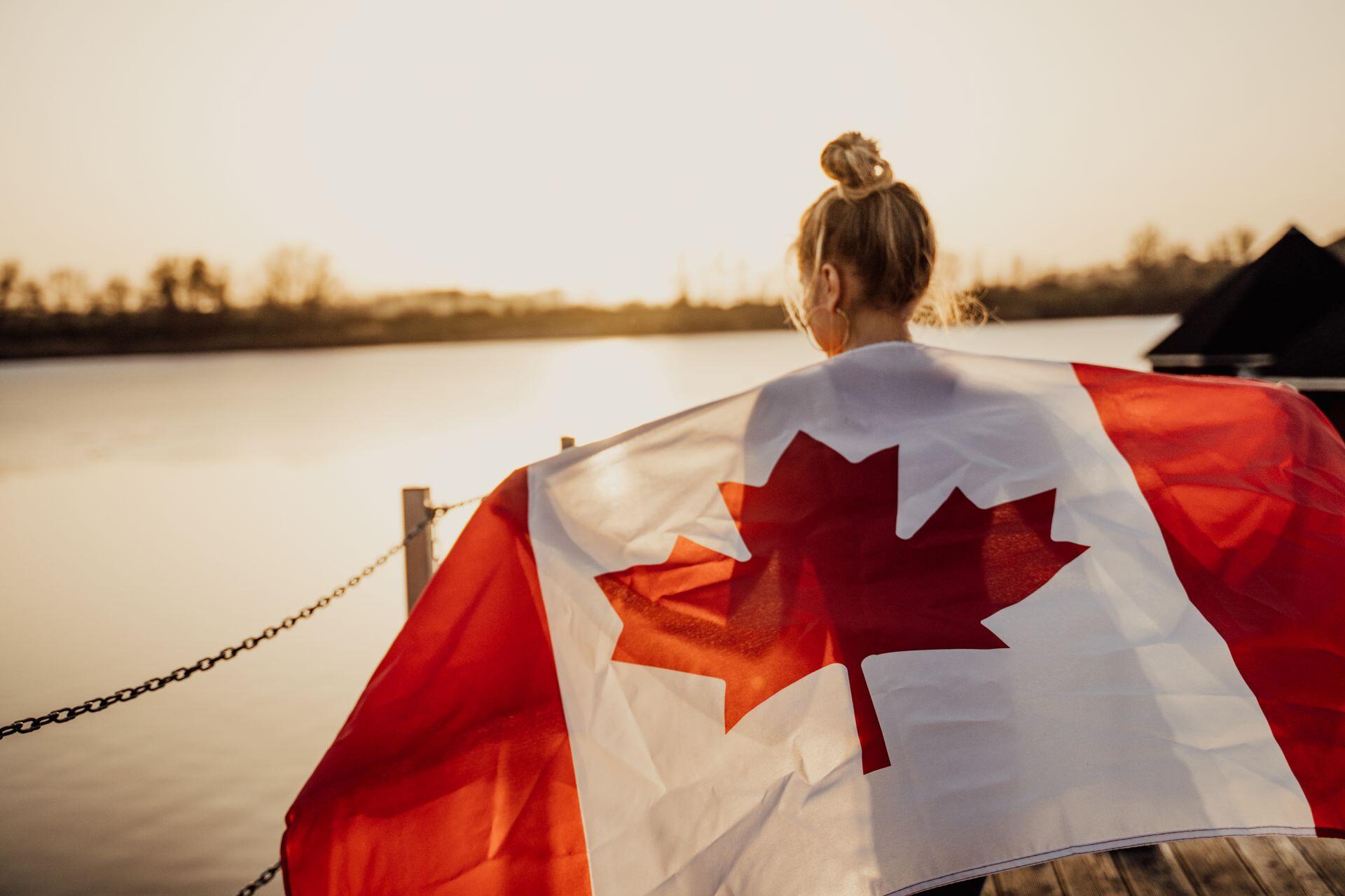 A girl wraps the Canadian flag around herself like a cape while standing in front of a sunset landscape with water and a far shore