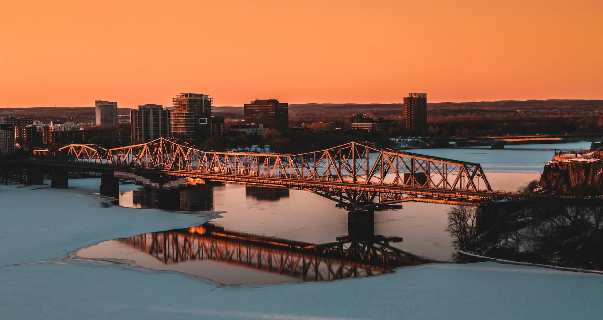 Orange sunset over a partially frozen river, with a steel truss bridge, city skyline, and reflections in the water.