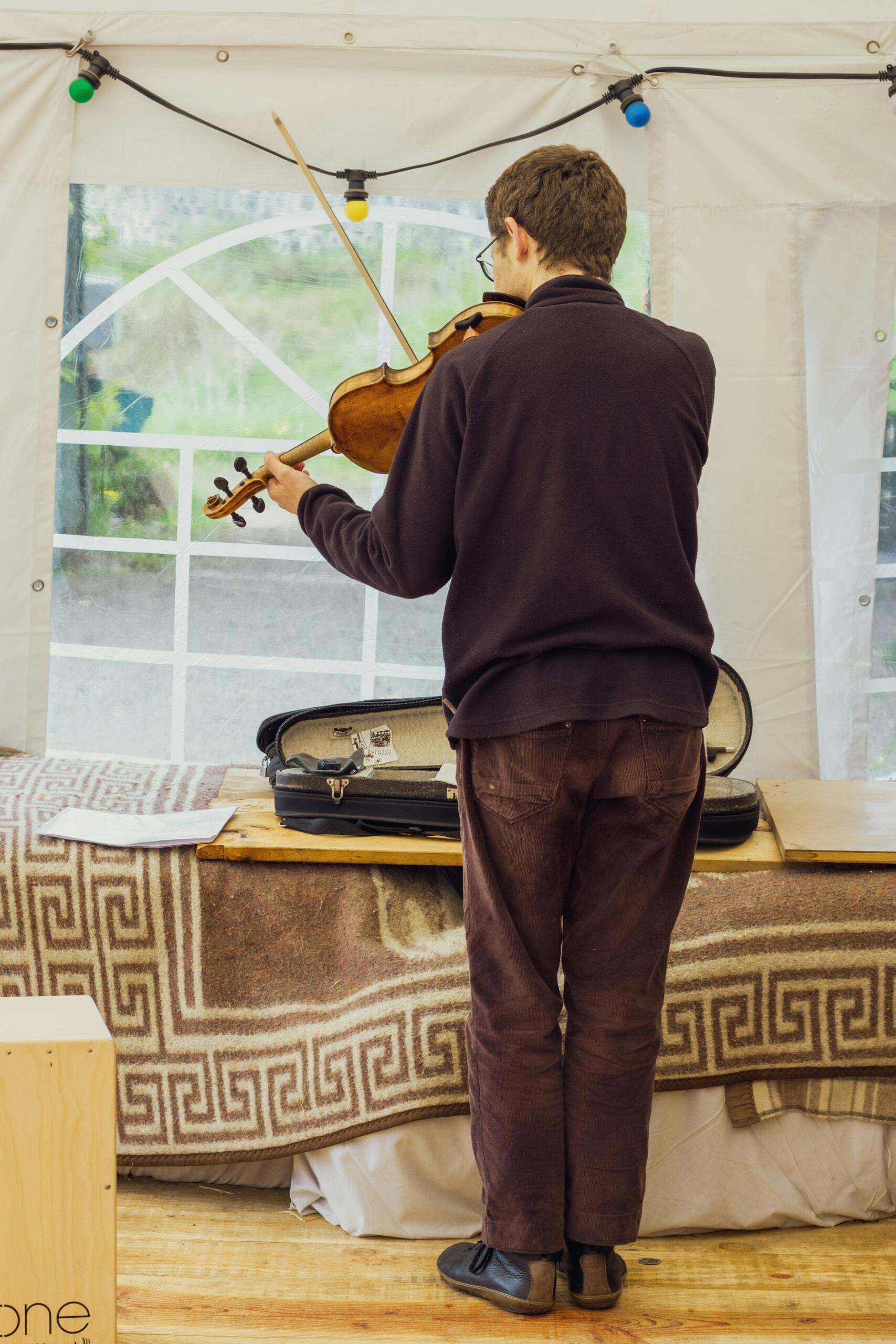 A musician plays violin inside a decorated tent, with colorful string lights and an open instrument case nearby.