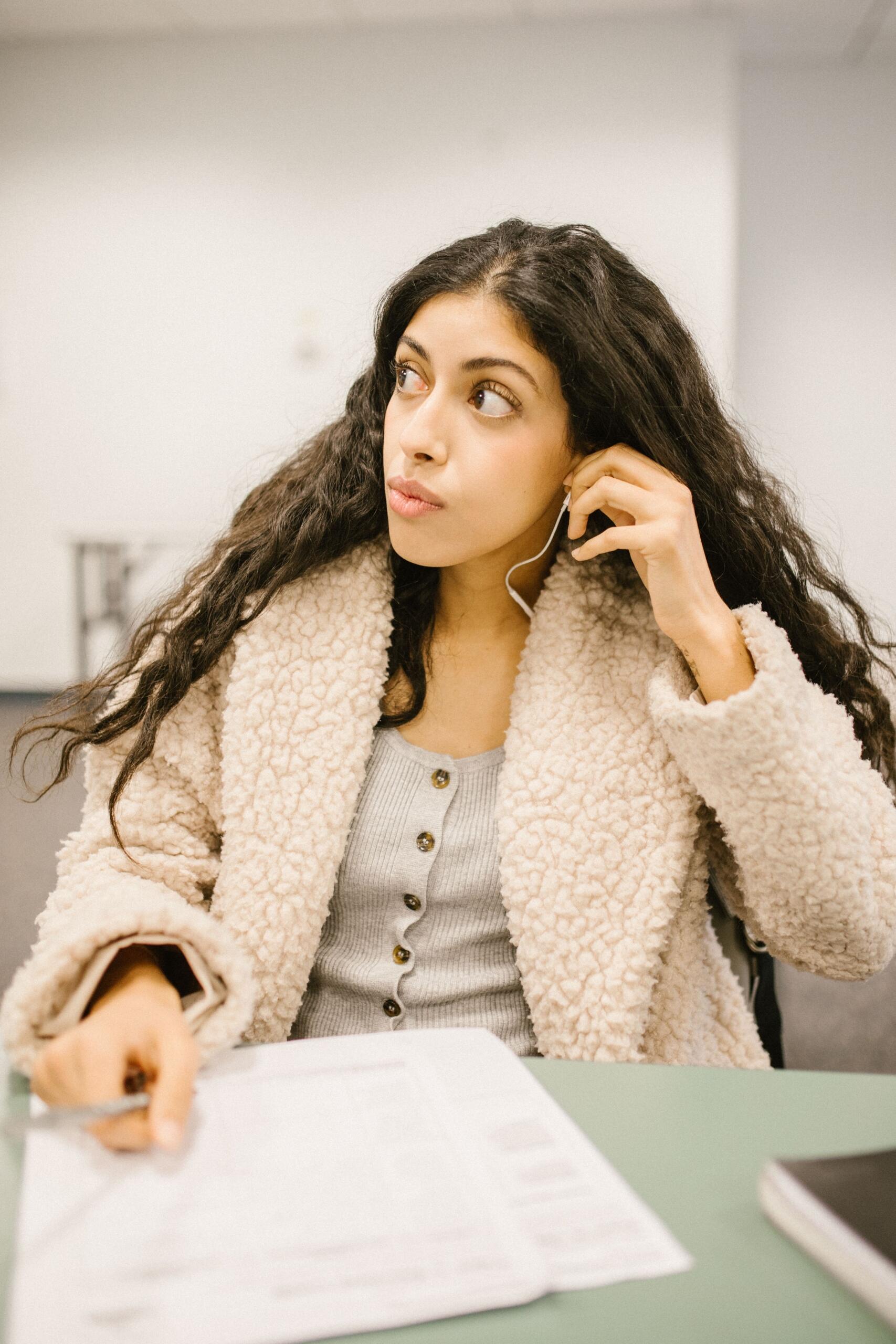 a young woman sits in a class, her work in front of her. She looks to the side, holding a headphone.