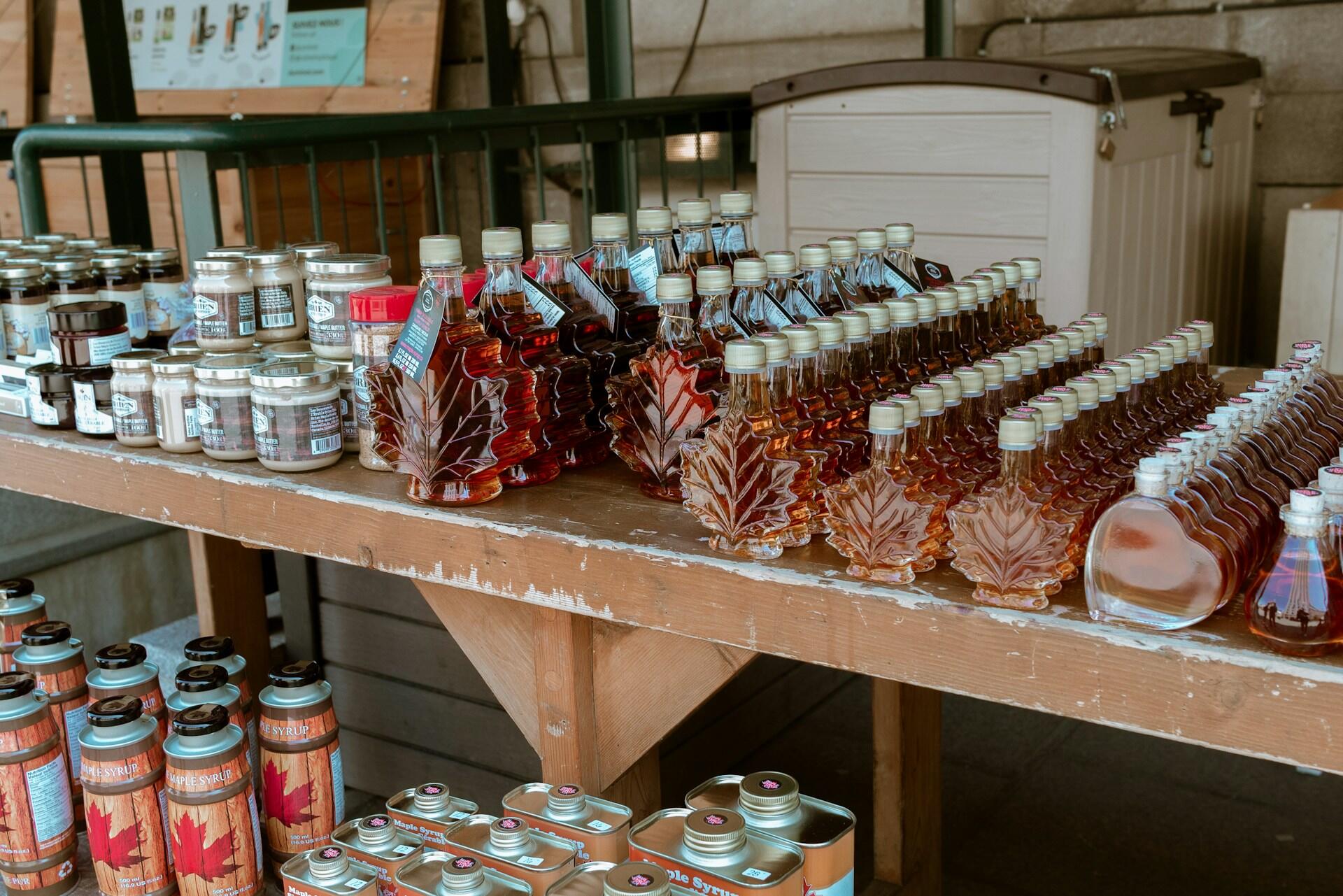 A market stall displays maple syrup in leaf-shaped bottles, jars, tins, and cans on wooden shelves.
