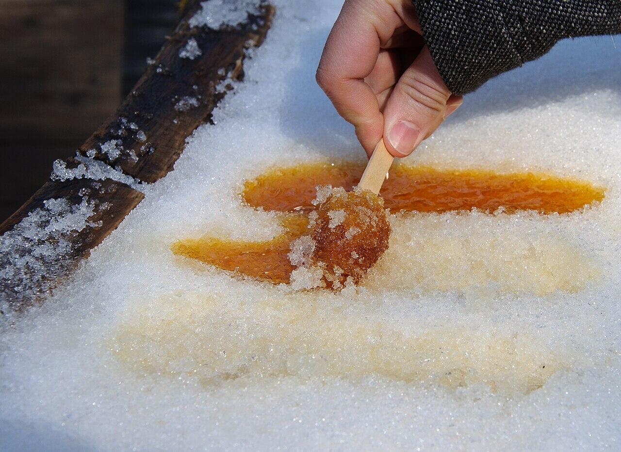 Hand holding a wooden stick collecting amber maple syrup candy freezing on snow outdoors.