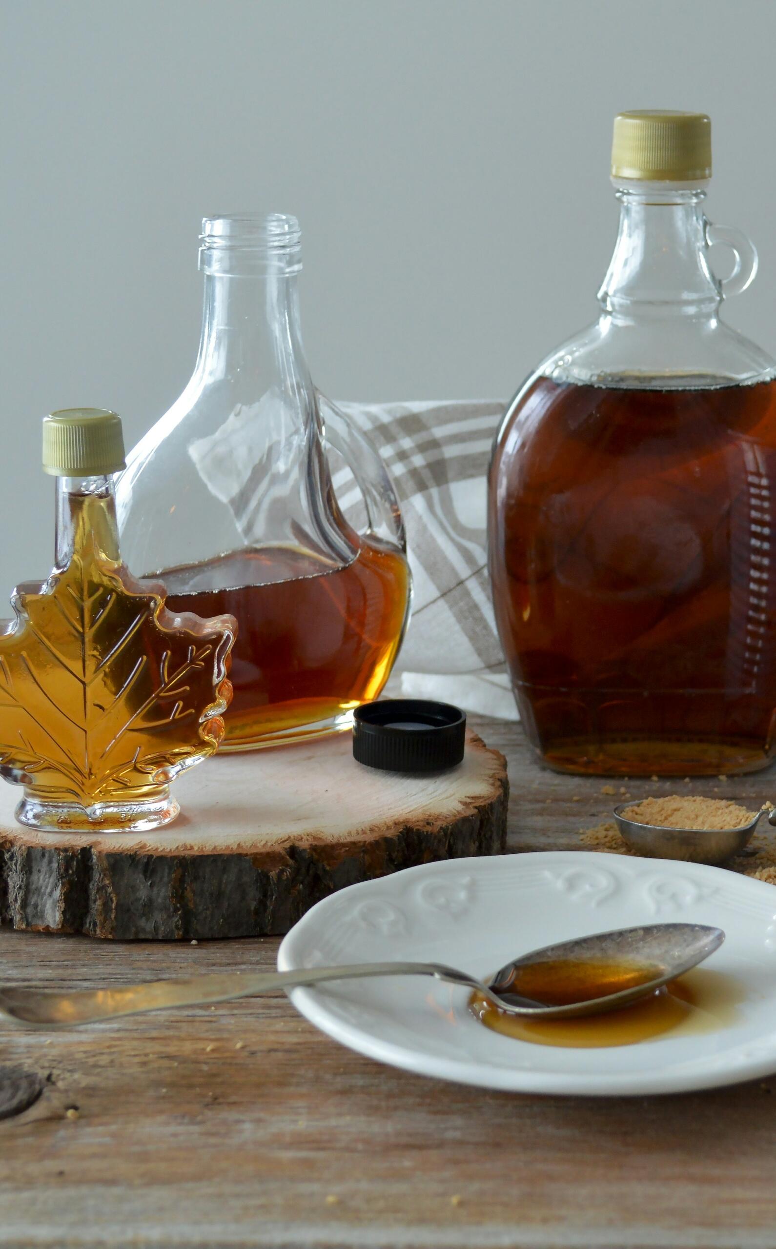Bottles of maple syrup, one shaped like a maple leaf, on a wooden surface with a spoon and syrup on a white plate.