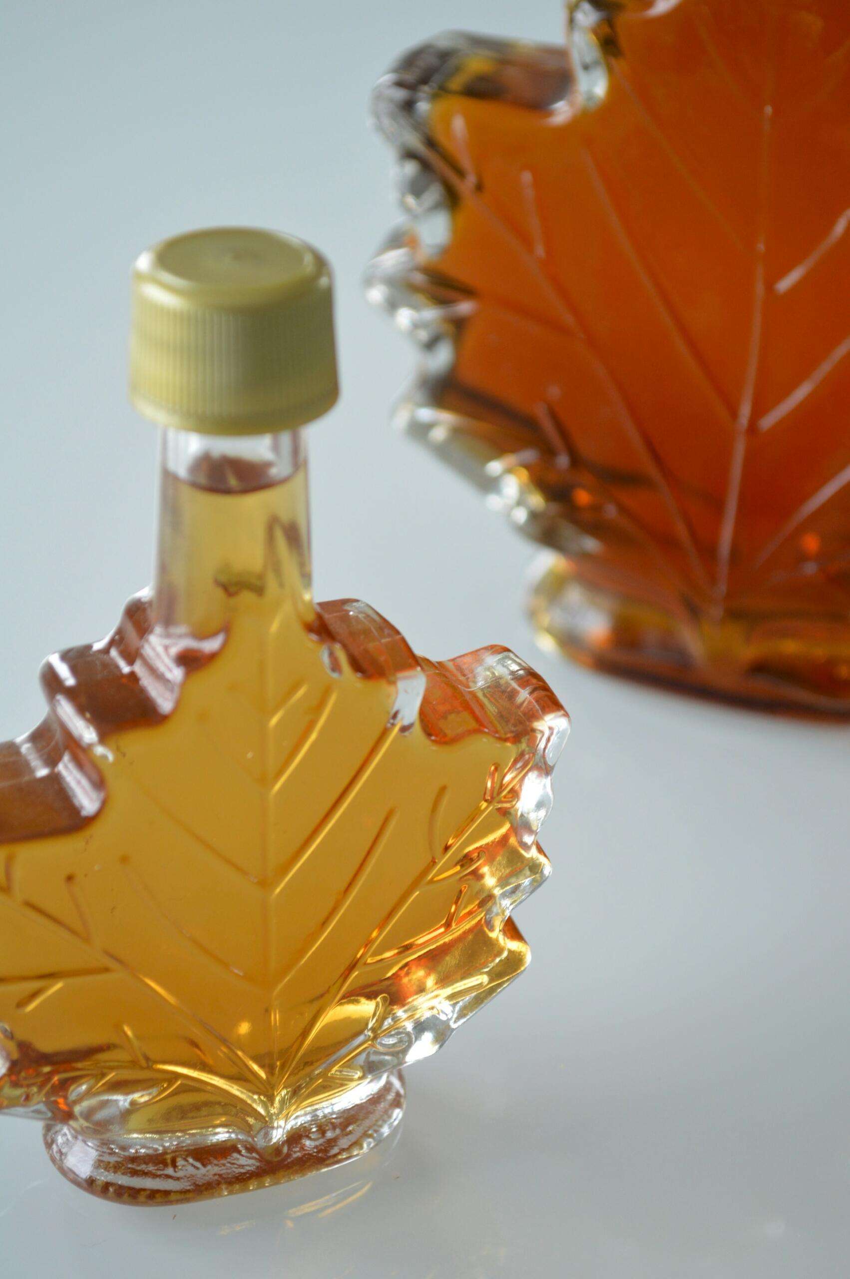Two glass maple leaf-shaped bottles filled with amber-colored maple syrup on a white surface.