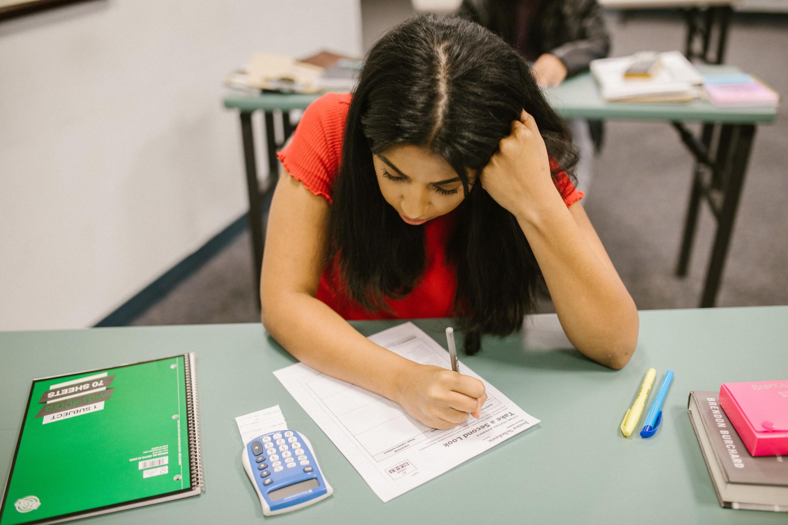 A teenage girl works at her desk. A green notebook and other school supplies are strewn across the desk.