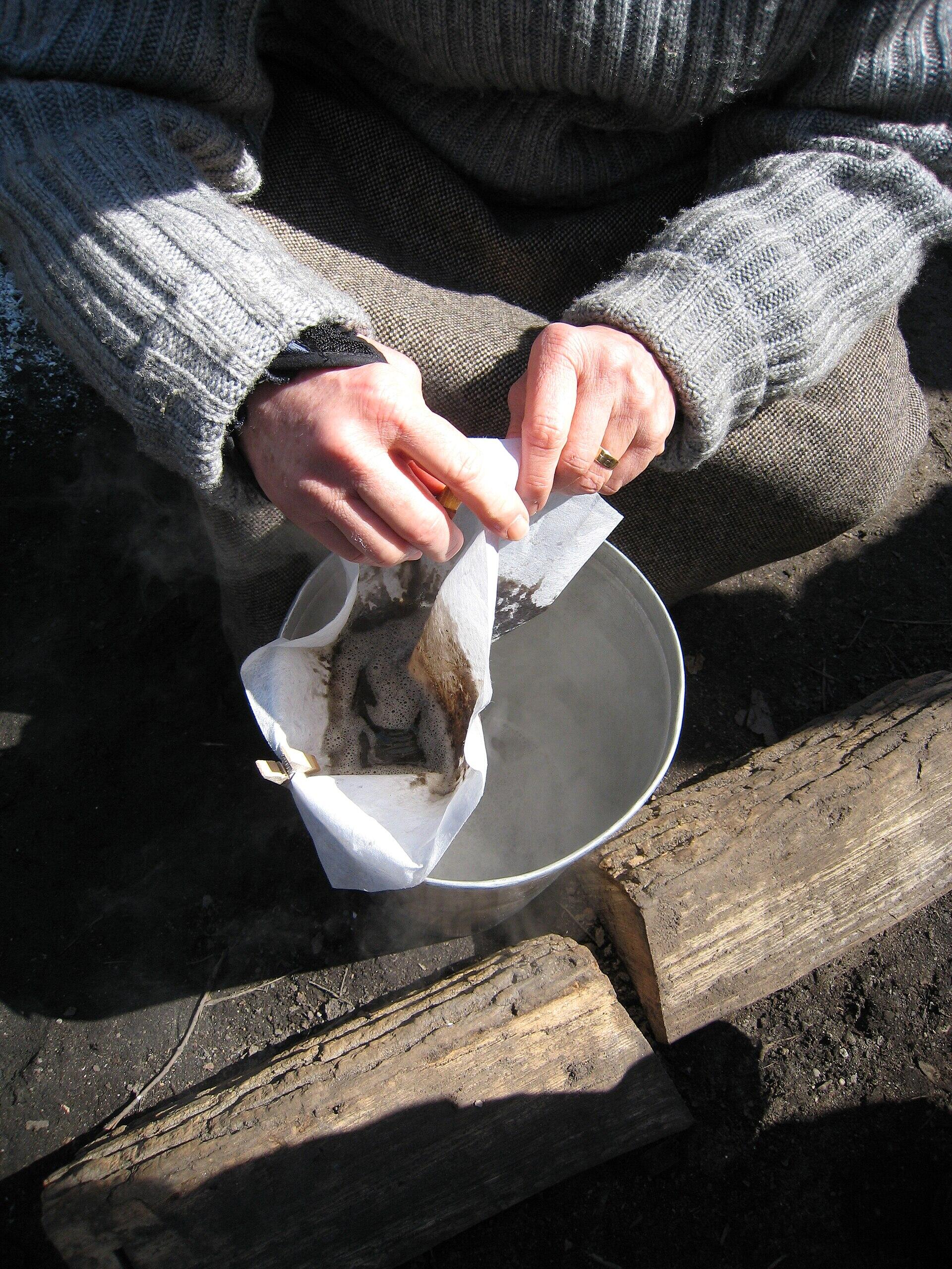 Person wearing a gray sweater brewing coffee outdoors using a cloth filter over a steaming metal pot with wooden logs nearby.