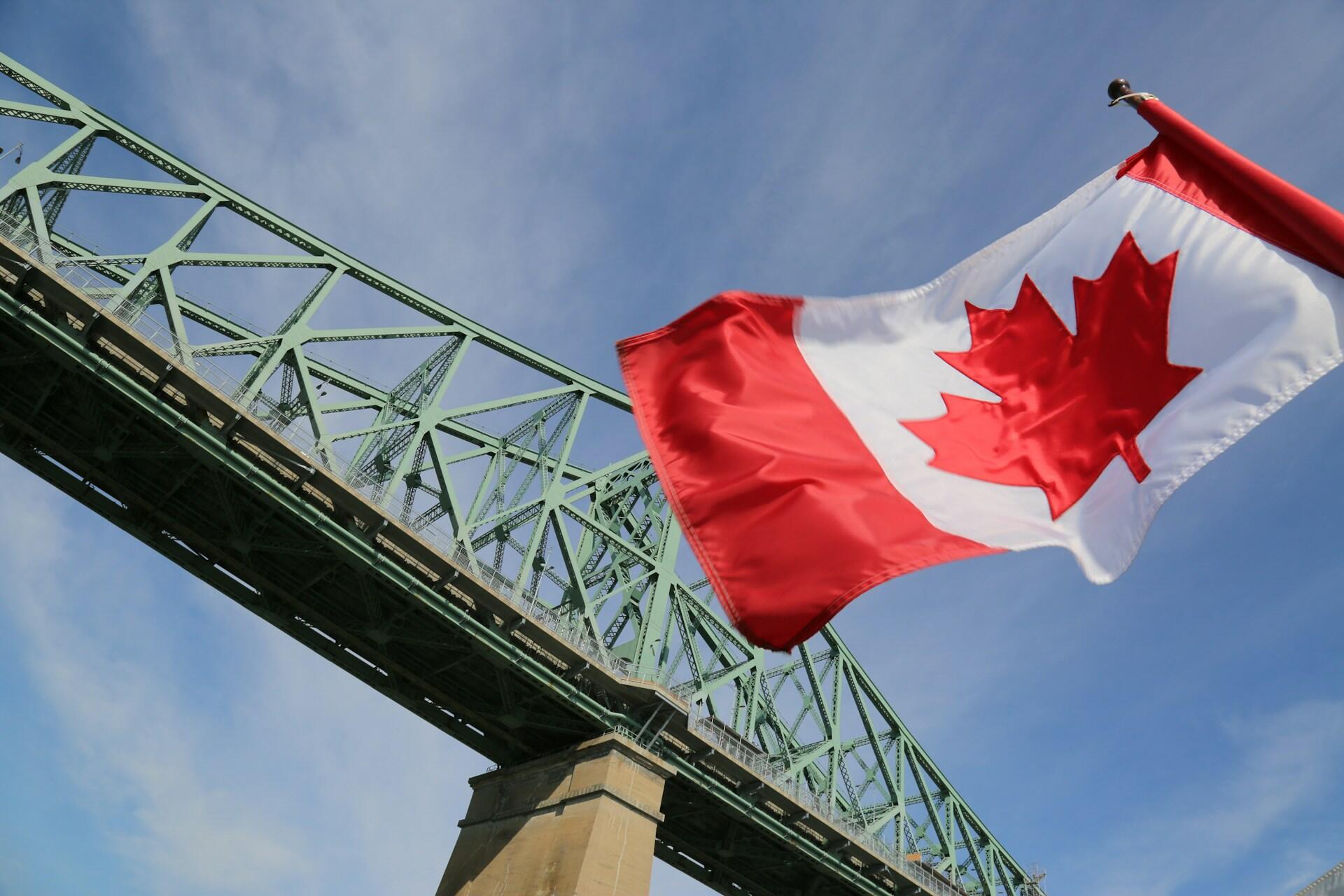 Canadian flag waving in front of a green steel truss bridge under a blue sky.