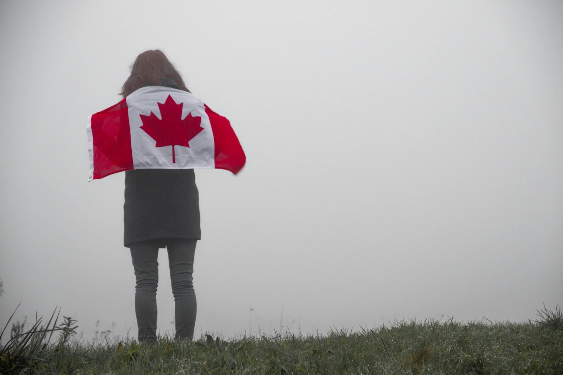 Person seen from behind holding a Canadian flag on a foggy grassy hill.