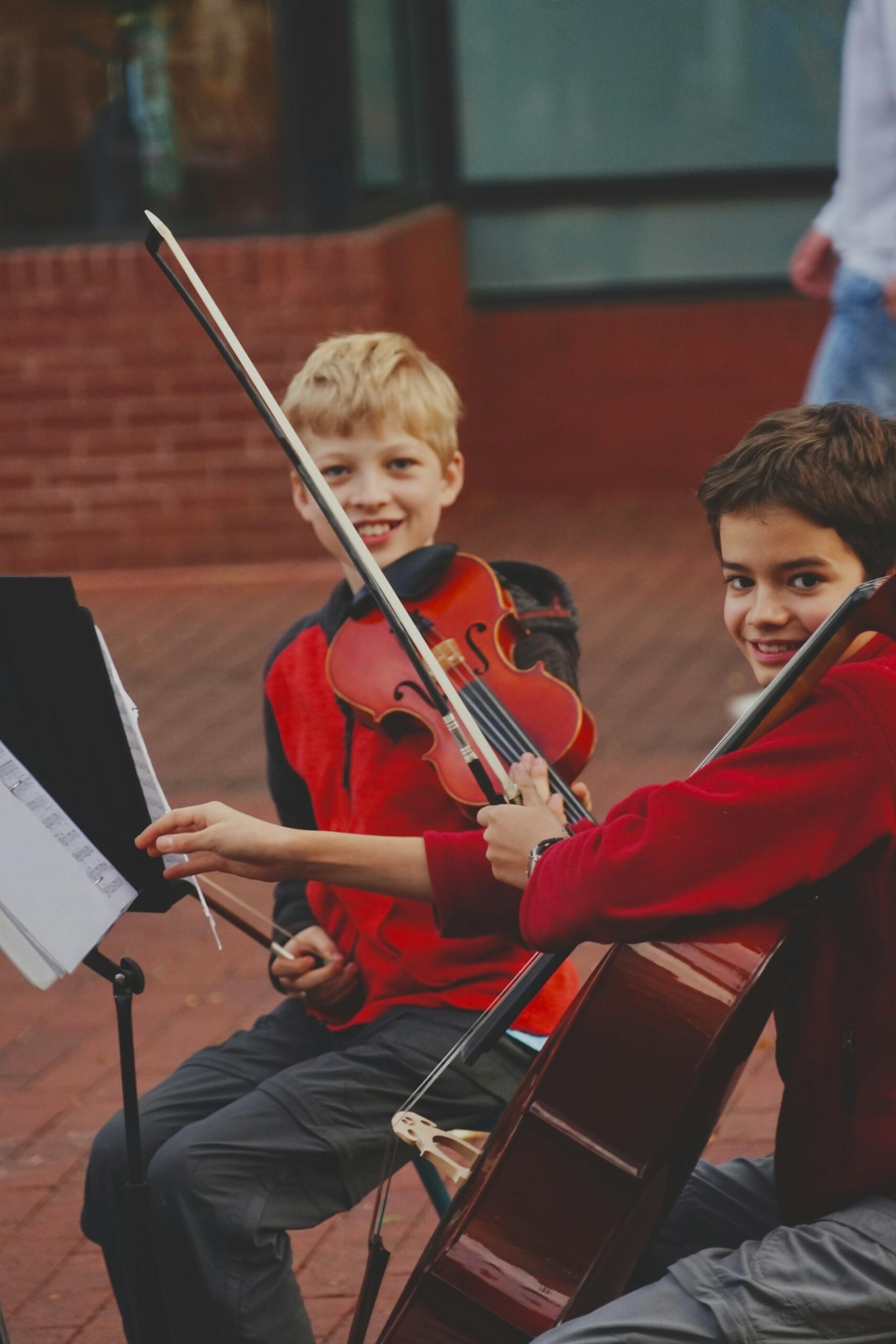 Two smiling children in red play string instruments together, with sheet music on a stand and a cello in the foreground.