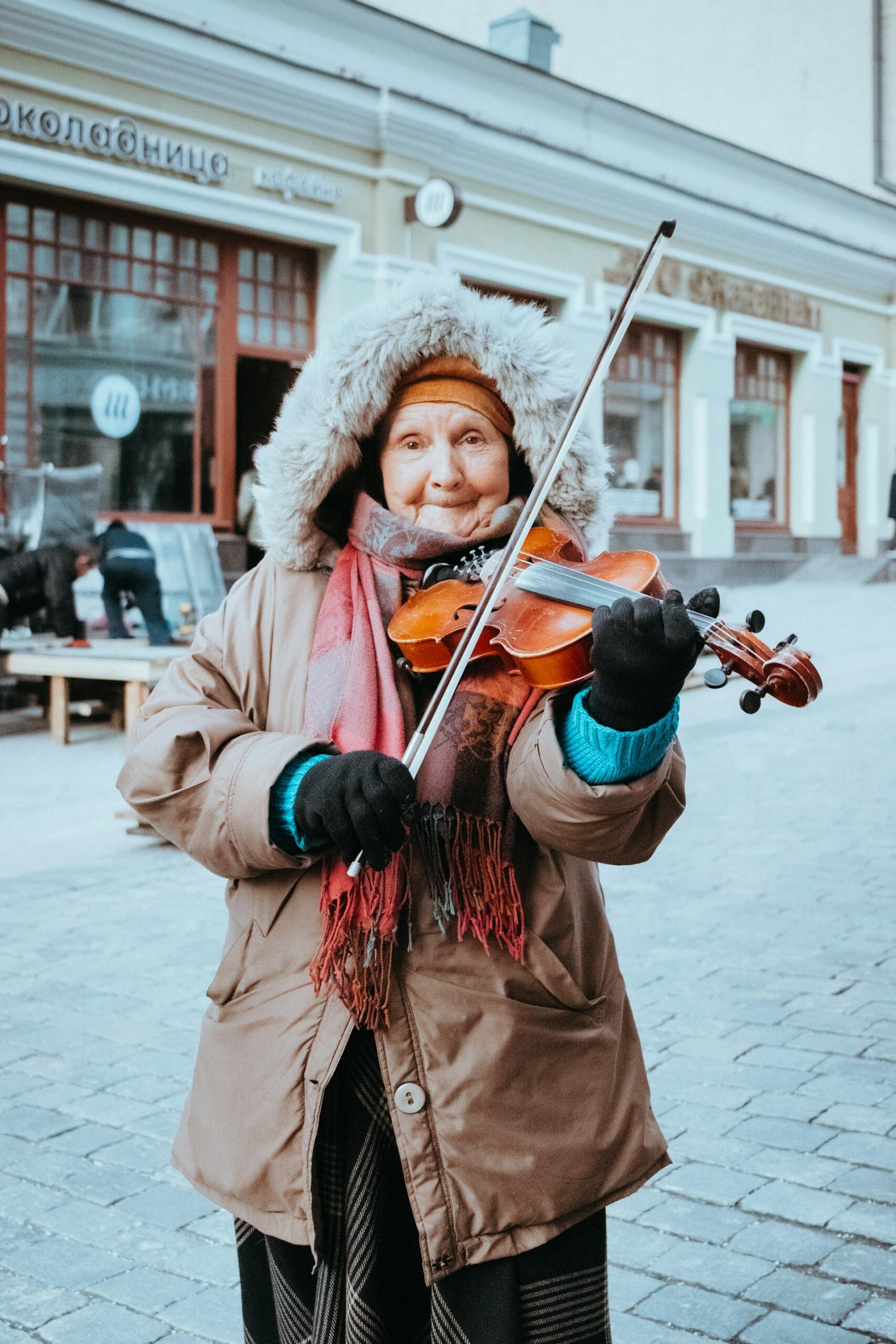 Street musician in winter clothing plays a violin outdoors in front of a building.