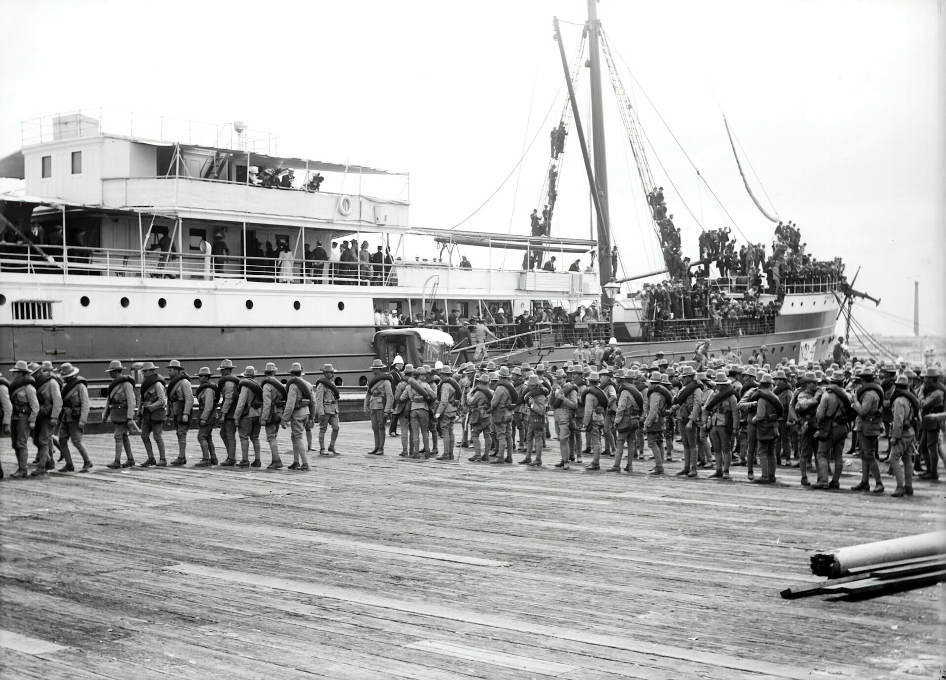 Black-and-white photo of soldiers boarding a crowded ship at a harbor, with troops lining the dock and climbing the vessel.