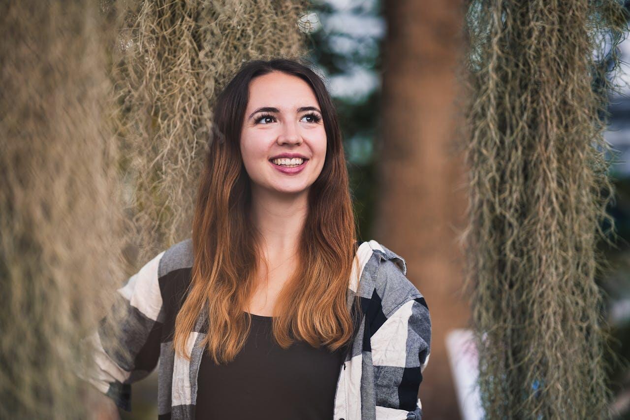 fille au cheveux bruns mi long souriant contre des arbres